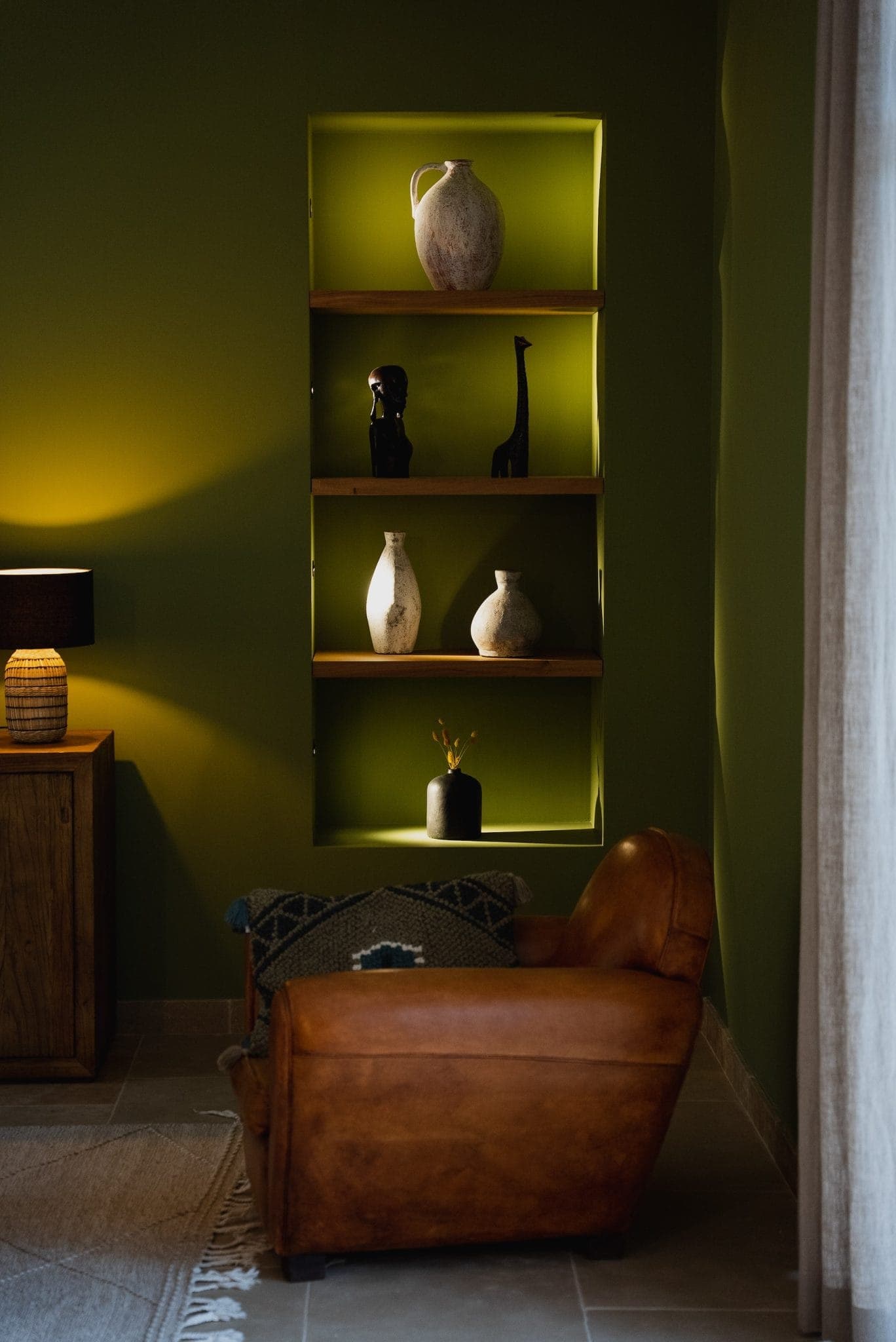 A warm room with green walls houses a leather armchair and a wooden side table with a lamp. Built-in shelves display decorative vases and animal figurines. Soft lighting enhances the warm, welcoming atmosphere.