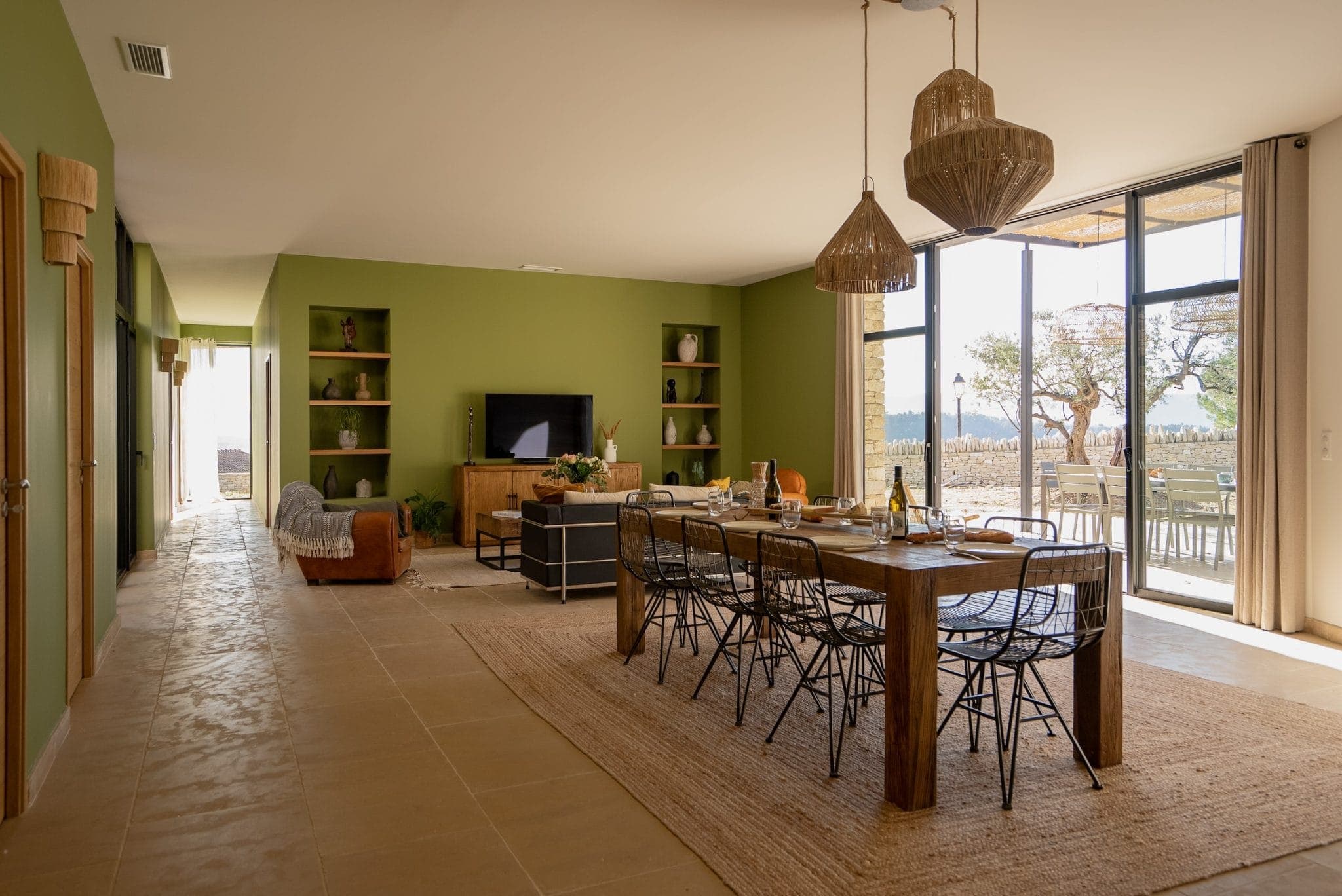 A spacious dining room with a wooden table for six, wicker hangings and large windows. The walls are olive green and the room includes a TV, shelving and a comfortable sitting area with views of the trees outside.