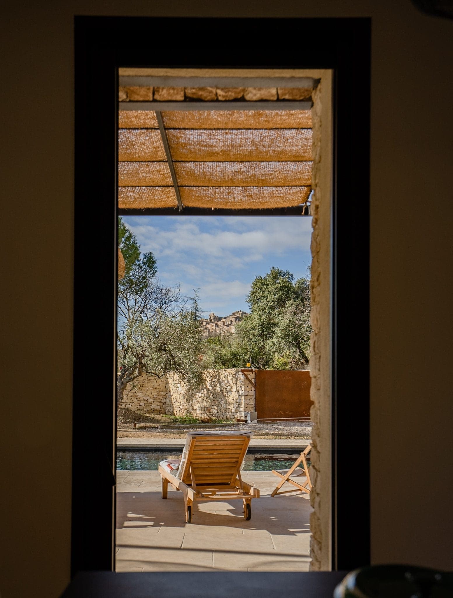 View through a front door of a sunny patio featuring a pair of wooden deckchairs facing a swimming pool, surrounded by a stone wall and trees under a clear blue sky.