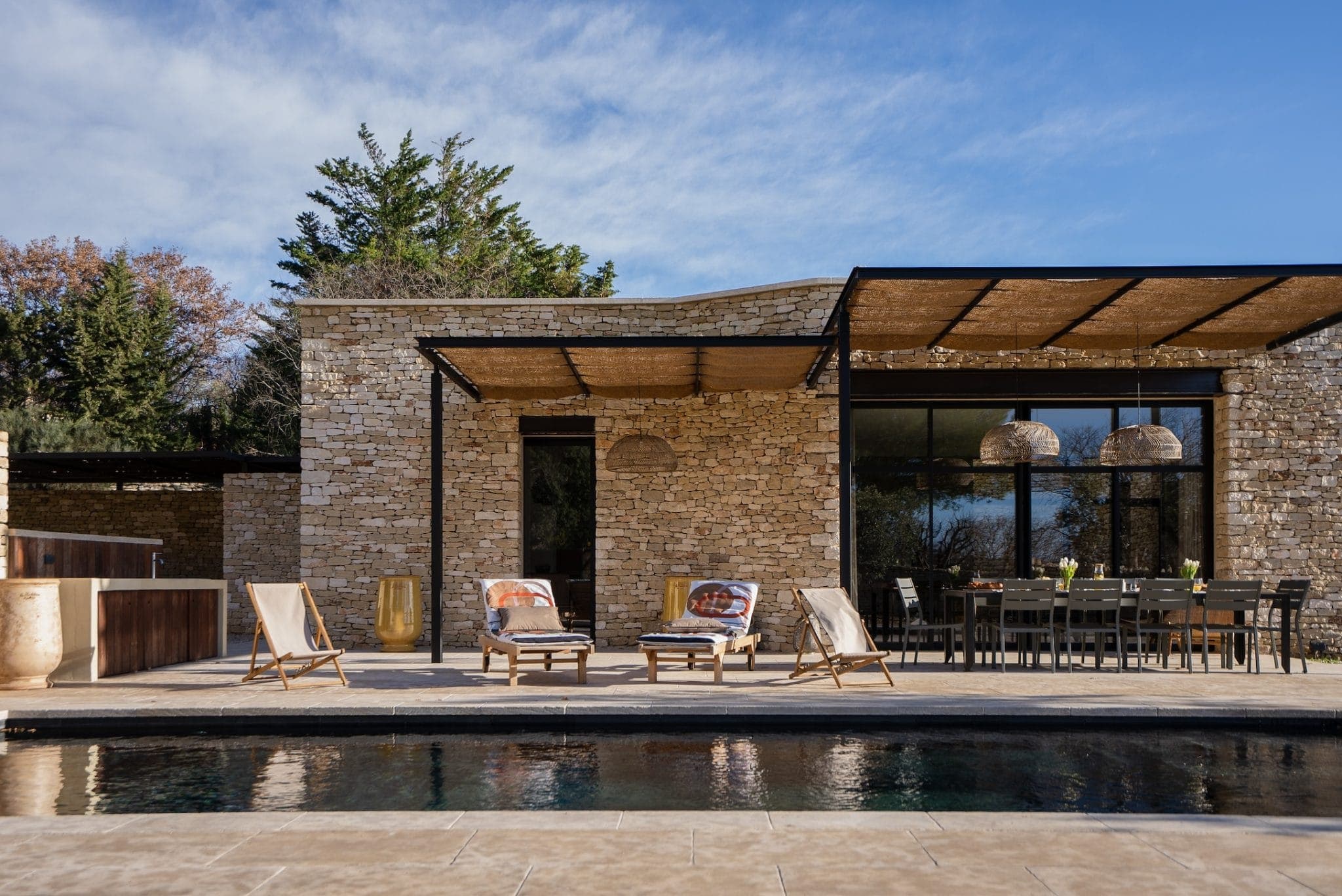 A modern stone house with a terrace equipped with deckchairs, a dining table and a swimming pool in the foreground. The sky is blue with light clouds and there are trees in the background.