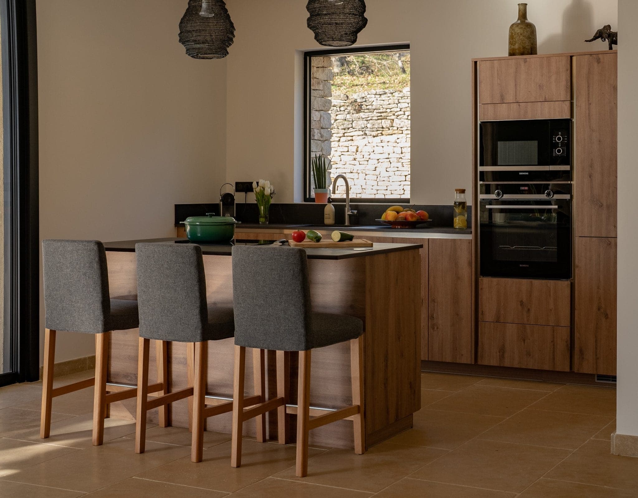 Modern kitchen with wooden island and gray chairs. Appliances include oven and microwave. The worktop houses a green saucepan, a cutting board with vegetables and a fruit bowl. Hanging lamps and a stone wall can be seen in the background.