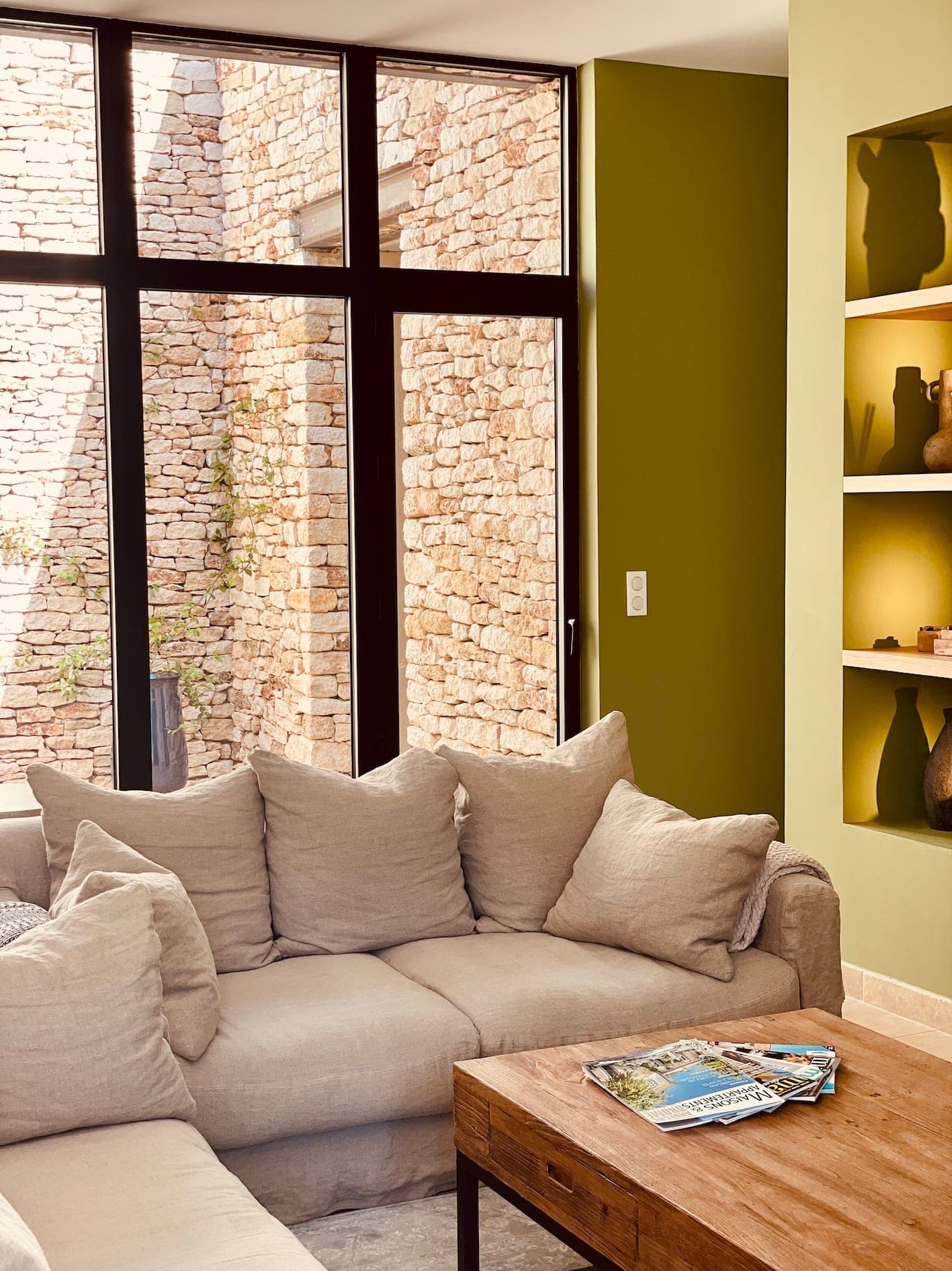A comfortable living room with a beige corner sofa with cushions. Sunlight streams through high windows, illuminating the exposed brick walls. A wooden coffee table holds magazines, while built-in shelves display a variety of decorative objects.