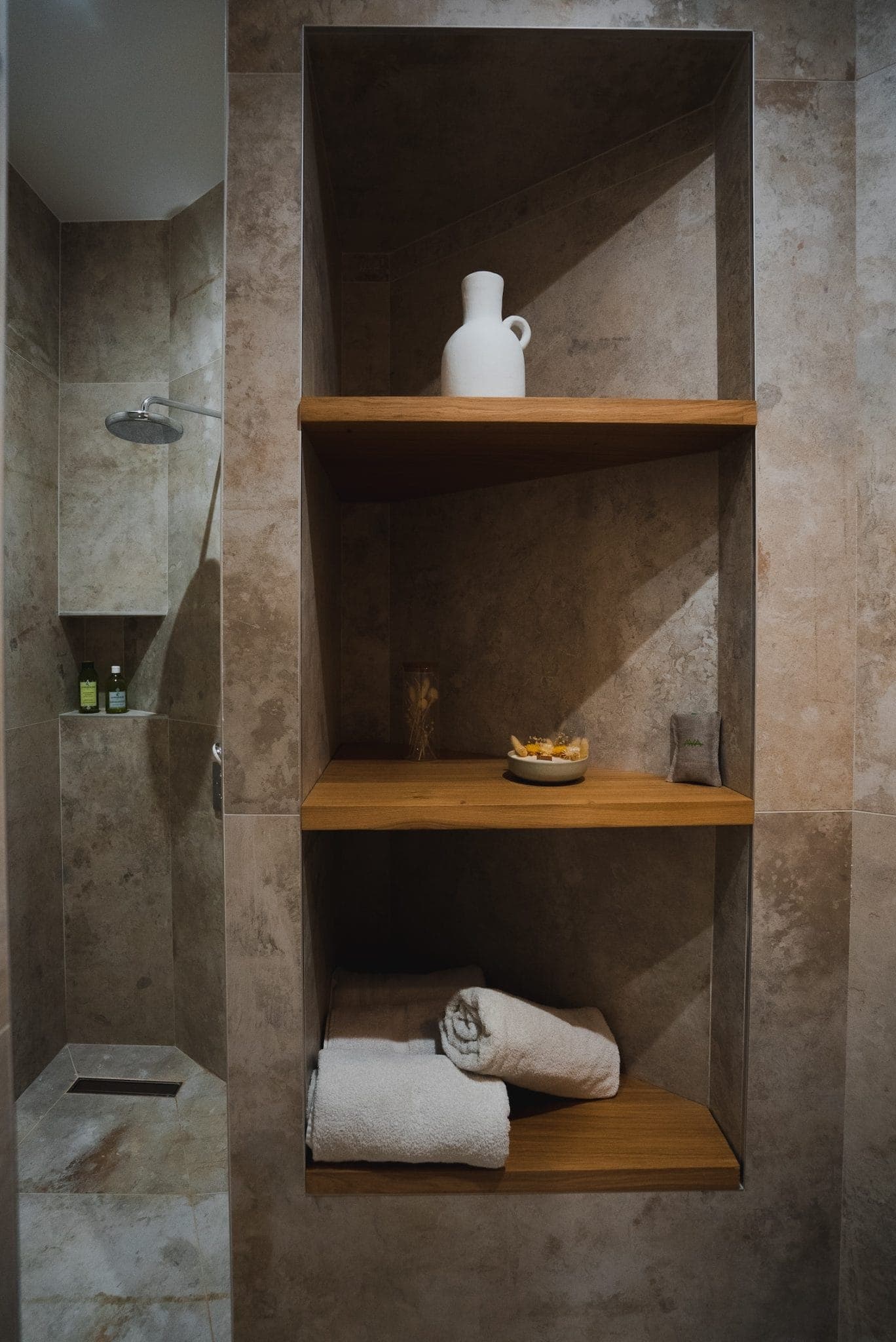 A minimalist bathroom with a polished stone shower featuring a chrome showerhead and two small bottles of shampoo. Adjacent wooden shelves hold a white carafe, a bowl with candles, glass vases and neatly folded towels.