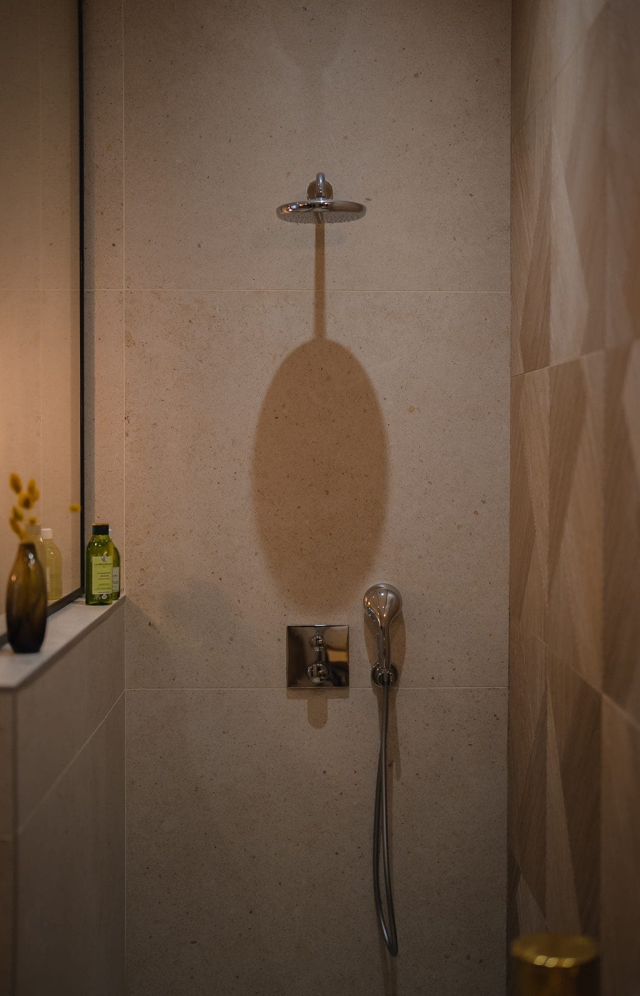 A minimalist bathroom with a wall-mounted shower head and a hand-held shower. The wall is tiled in beige and there's a shelf with a yellow vase and a green bottle. The lighting is soft and warm, creating a cosy atmosphere.