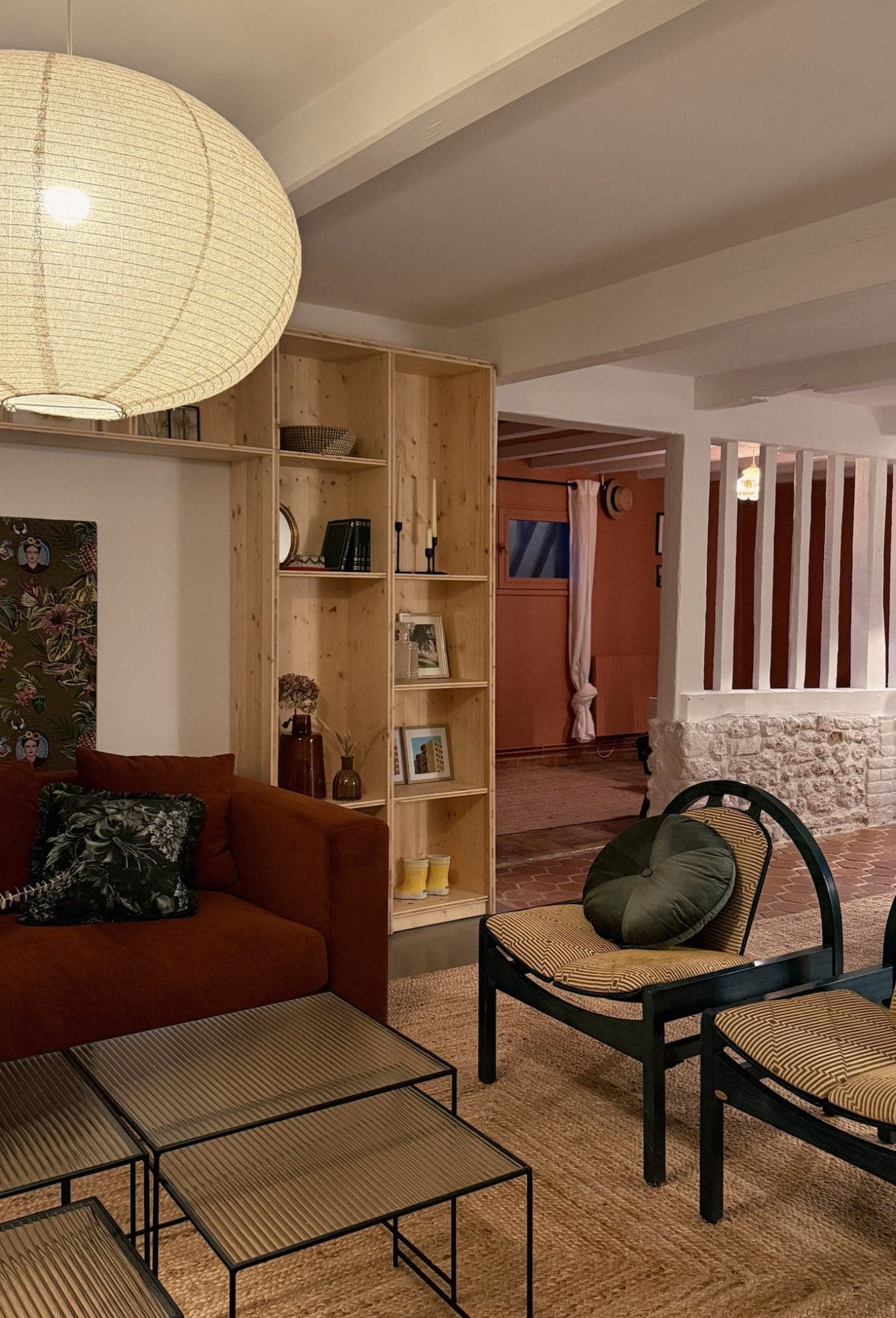 Un salon confortable avec un canapé couleur rouille, deux chaises rondes en osier et une table basse à plusieurs niveaux. Une grande lanterne en papier est suspendue au plafond. Une bibliothèque en bois avec des livres et des objets de décoration se trouve à l'arrière-plan.