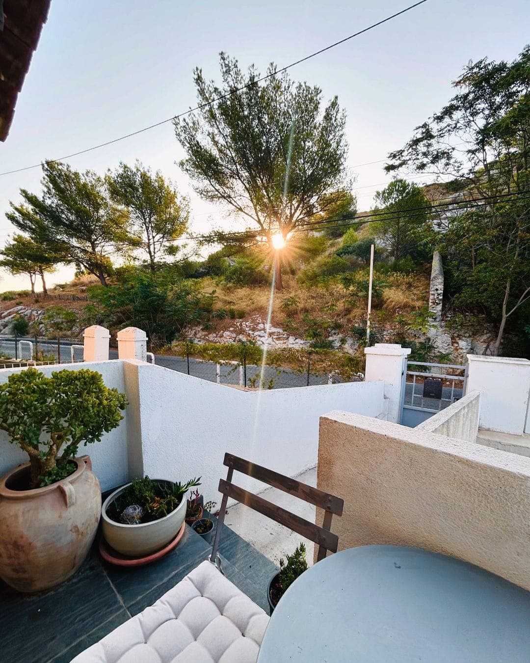 Un patio tranquille avec un banc rembourré et une table ronde est baigné de soleil. Des plantes en pot ornent l'espace, tandis qu'une vue panoramique sur les arbres et un soleil couchant créent une toile de fond paisible au-dessus d'une clôture blanche.
