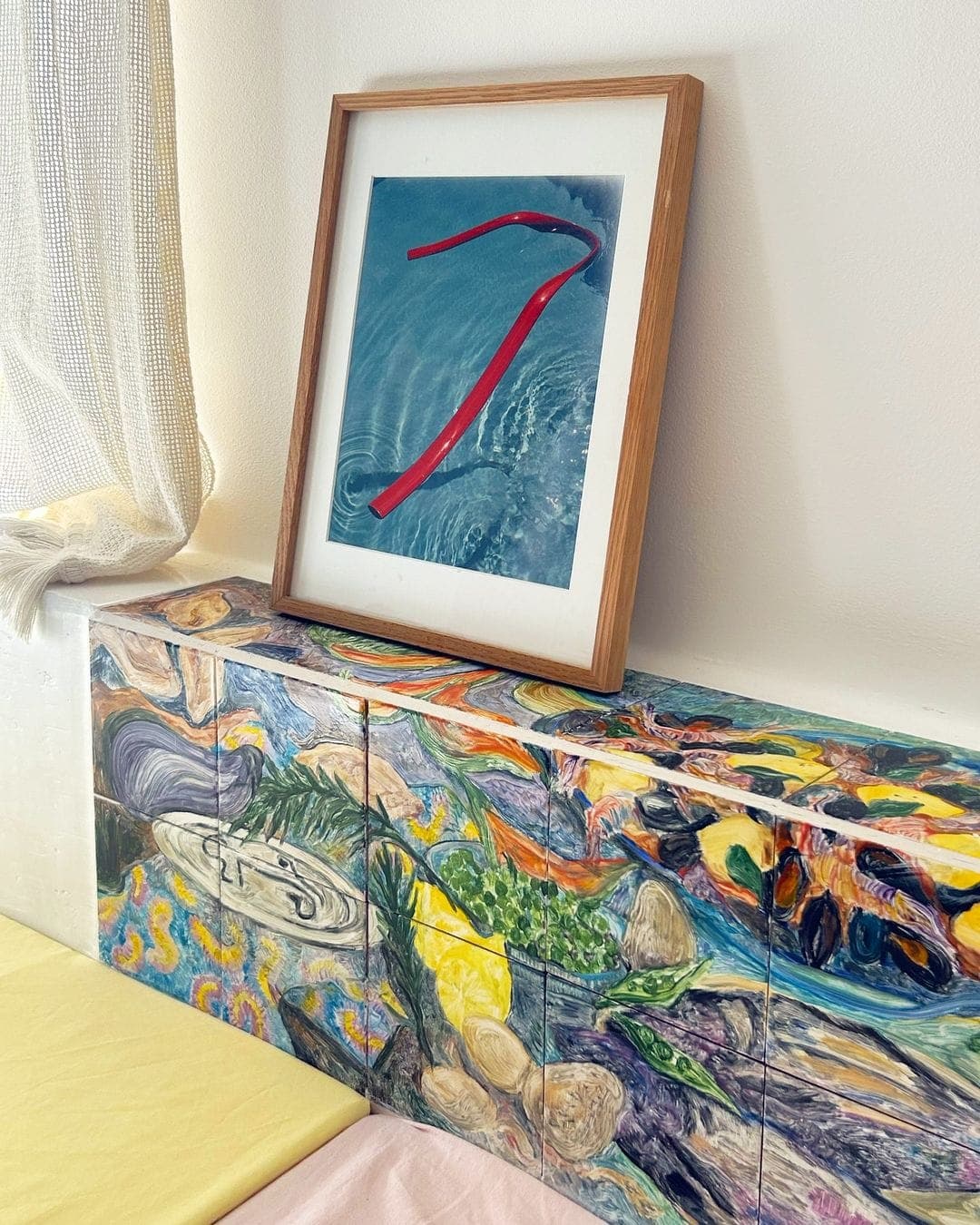 Une œuvre d'art abstraite encadrée avec une ligne courbe rouge sur fond bleu est appuyée contre un mur. En dessous, une surface colorée et texturée avec des motifs abstraits et mixtes ajoute de la vivacité. Un rideau léger se drape sur le côté.