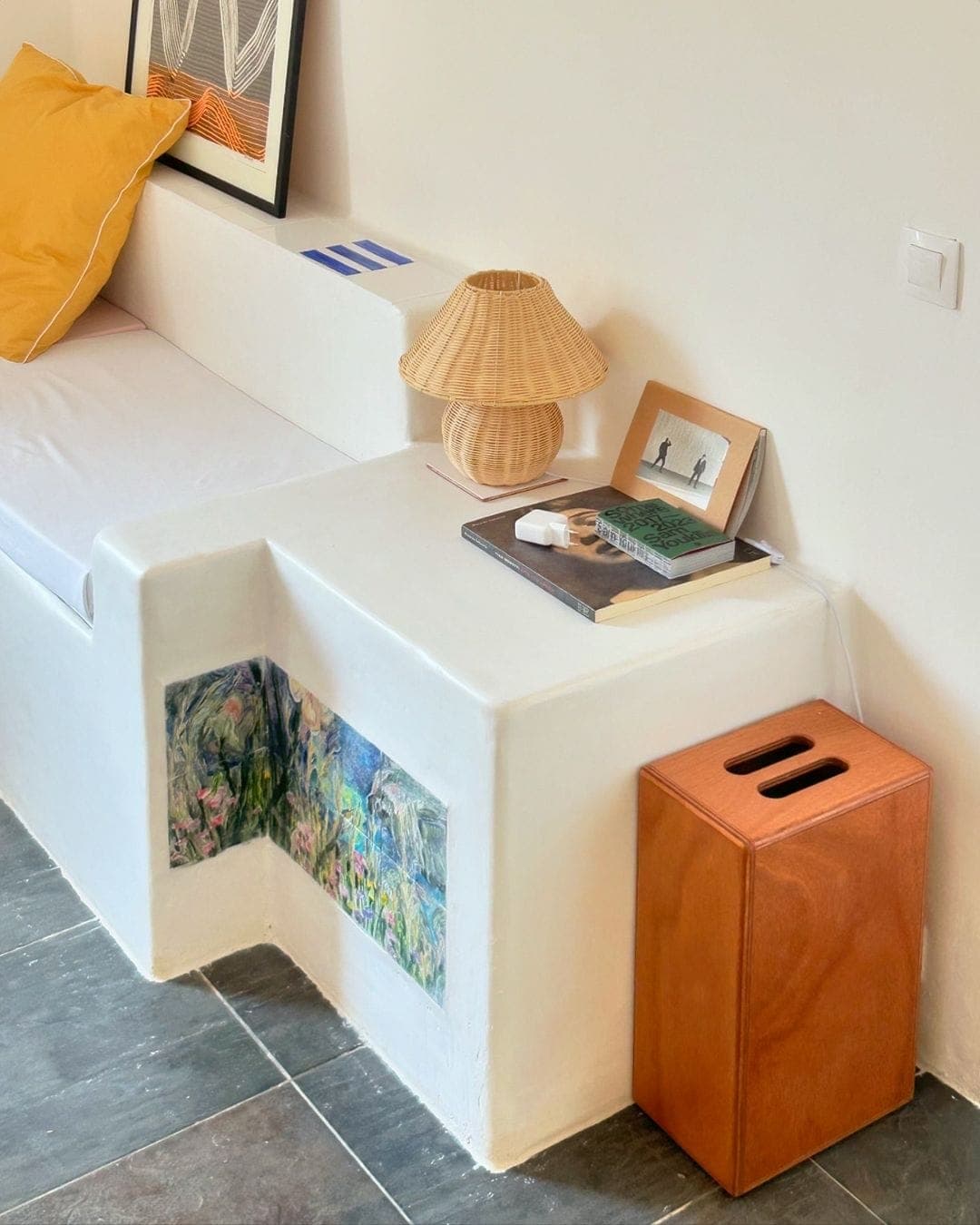 Un coin cosy comprend un coin salon blanc intégré avec des coussins jaunes. Une petite table y est fixée, sur laquelle sont posées une lampe en osier, des livres et des œuvres d'art encadrées. Une boîte en bois est posée sur le sol à proximité. Le mur comprend une décoration florale peinte.