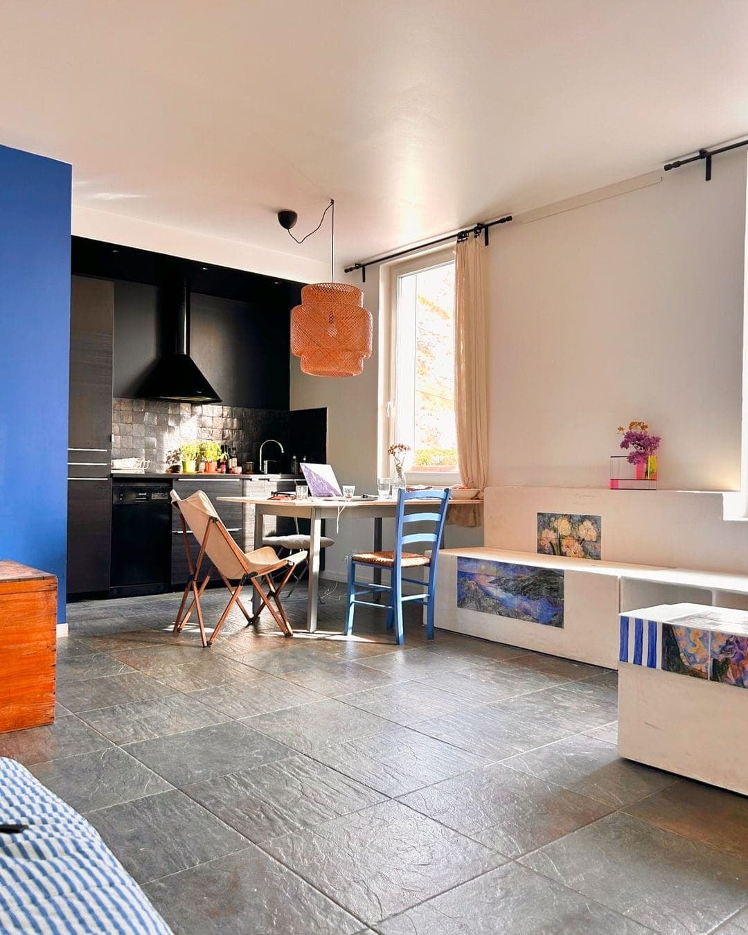 A cozy apartment with a modern kitchen, featuring a black backsplash and appliances. A small dining table with a laptop and chairs sits near a window. The living area has a mix of blue and white decor with slate flooring, and a pendant lamp hangs above.