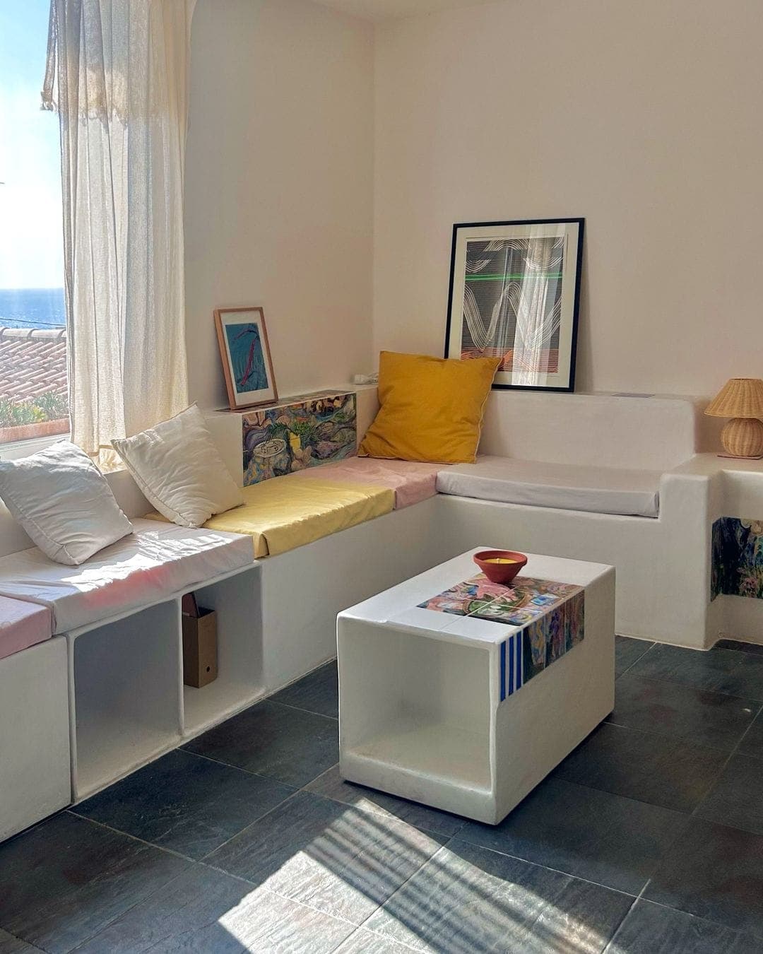Un salon chaleureux avec des sièges blancs intégrés ornés de coussins colorés. La lumière du soleil filtre à travers des rideaux transparents. Une table basse blanche et des œuvres d'art encadrées ajoutent au décor minimaliste, aux côtés d'une simple lampe sur le siège.
