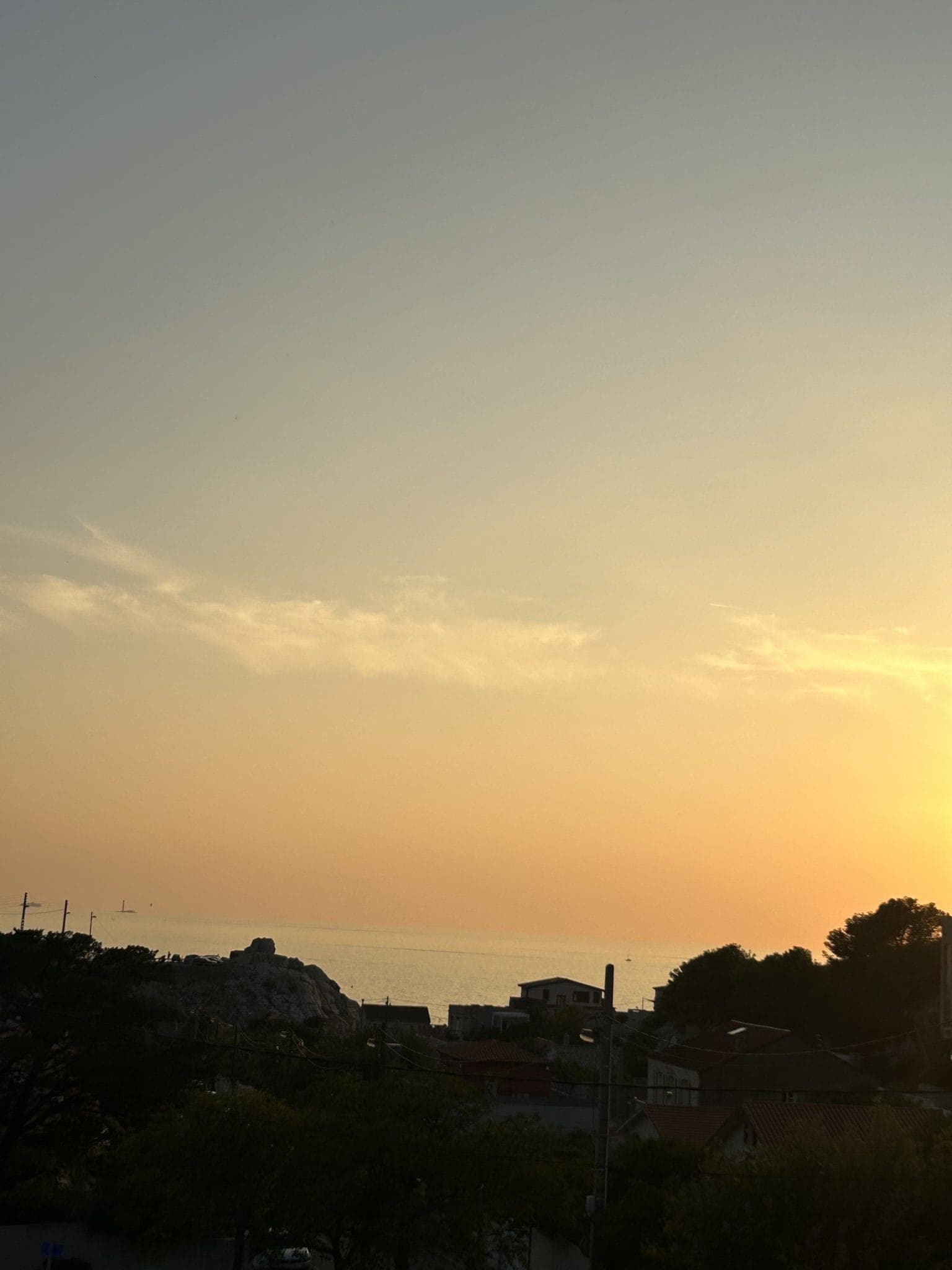 Un coucher de soleil serein sur un village côtier, avec un ciel orange et jaune doux se reflétant sur la mer calme. Les silhouettes des maisons, des arbres et des collines lointaines créent une scène tranquille et pittoresque.