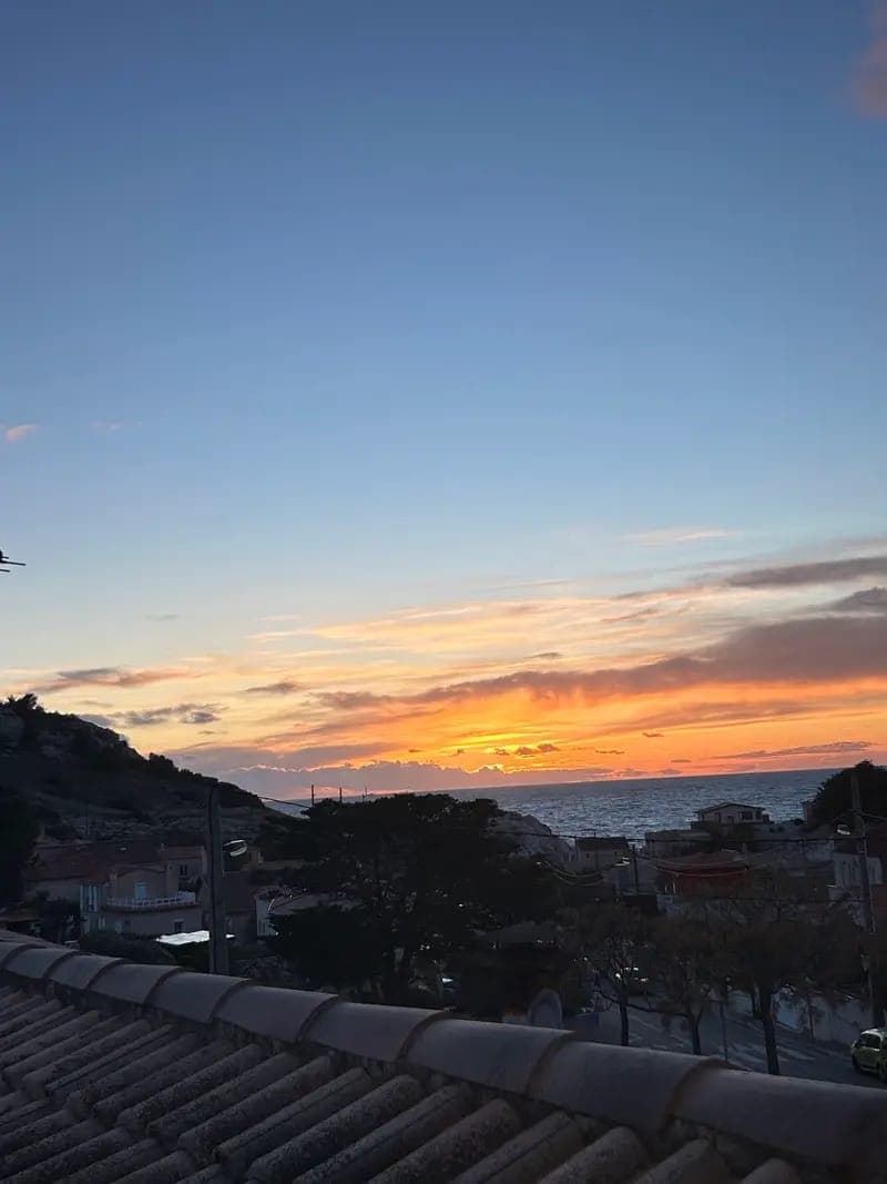 Vue panoramique d'une ville côtière au coucher du soleil, avec un ciel orange et jaune éclatant se fondant dans un bleu profond alors que le soleil descend derrière l'horizon. Les toits, les arbres et la mer sont visibles sous le ciel coloré du crépuscule.