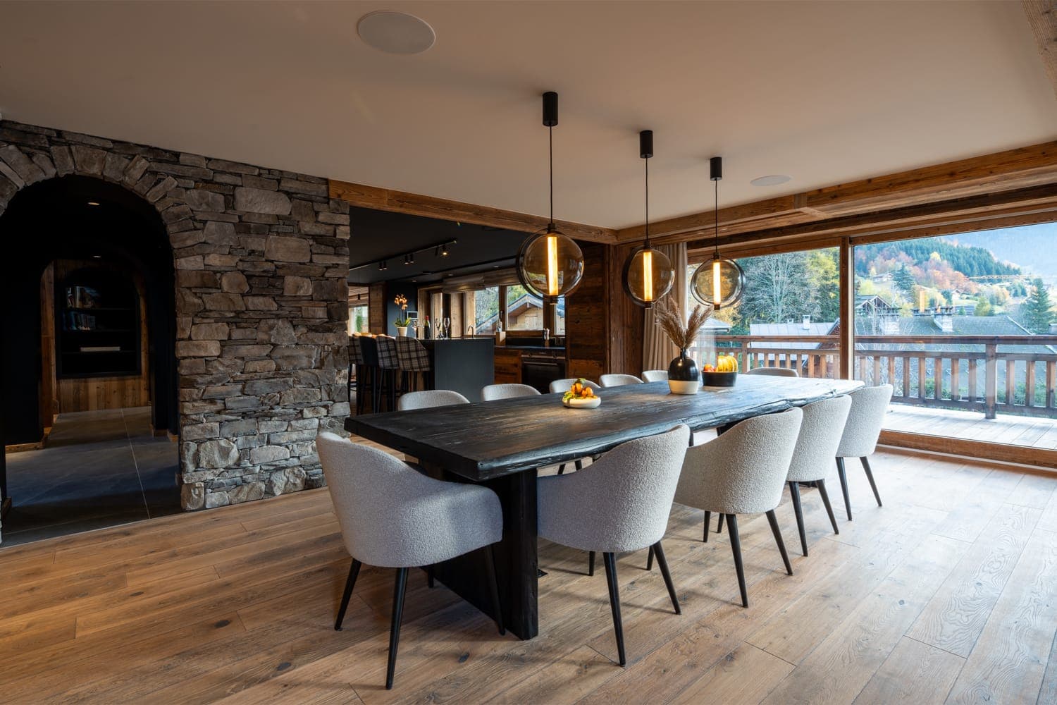 Une salle à manger spacieuse avec une grande table en bois entourée de chaises grises. Trois suspensions sont suspendues au-dessus. Un mur en pierre se trouve à gauche et de grandes fenêtres à droite offrent une vue sur la montagne et un aperçu d'un balcon.