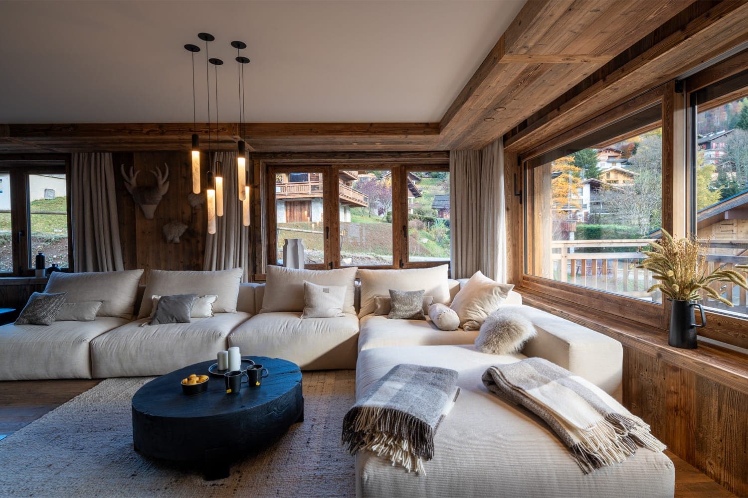A warm living room with large windows, a beige corner sofa covered with cushions and plaids, a round black coffee table with candles and wooden walls. Outside, wooden cabins and greenery are visible. The space exudes a rustic, welcoming atmosphere.