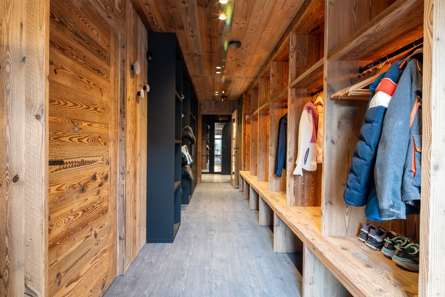Un couloir aux murs et au plafond en bois, avec des casiers ouverts remplis de vestes. Plusieurs paires de chaussures sont posées sur le sol. La lumière naturelle illumine l'espace, lui donnant une atmosphère chaleureuse et rustique.