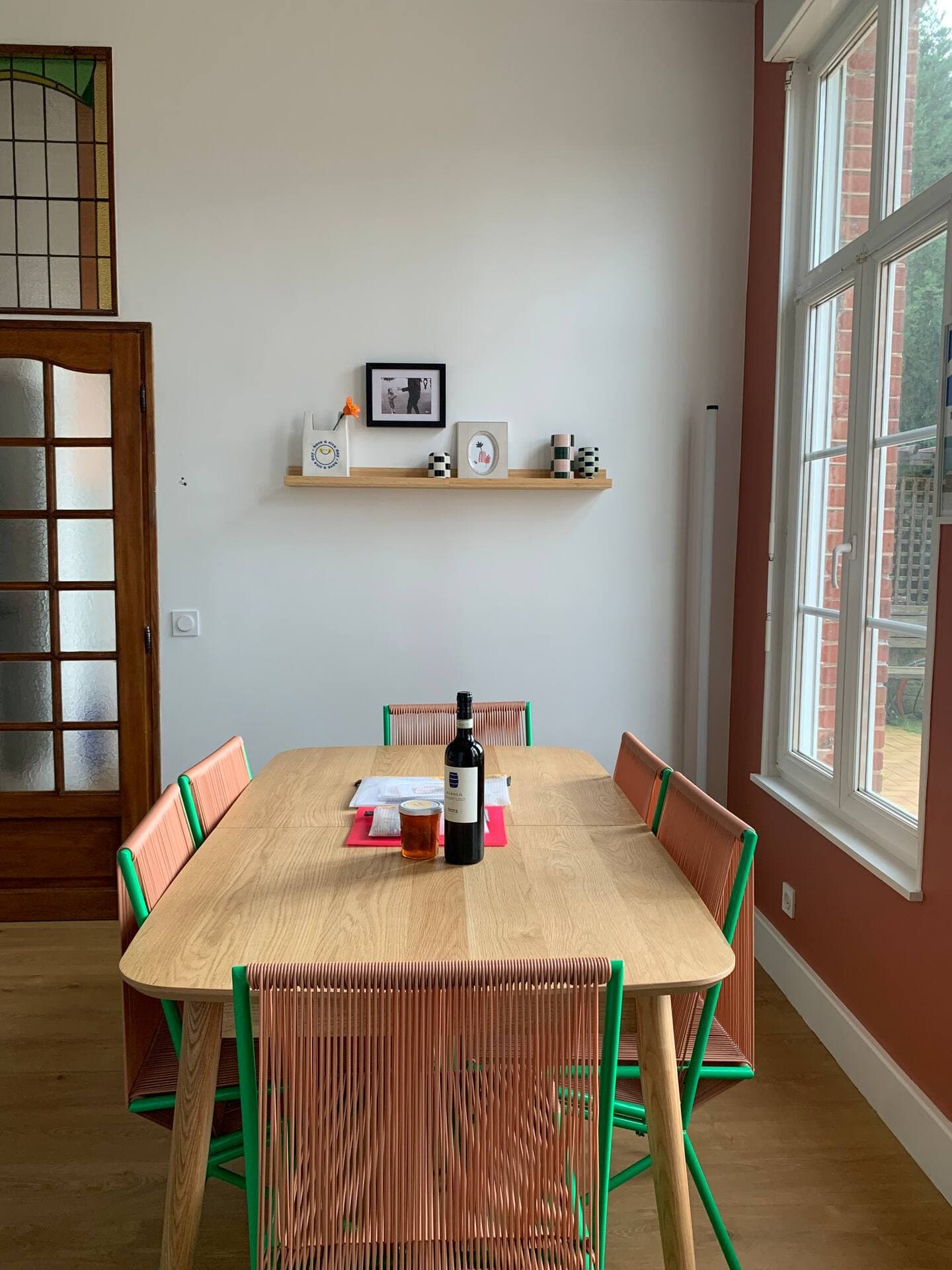 A dining room with a wooden table and six colorful chairs. A bottle of wine and a candle are on the table. A minimalist wall shelf holds decor items and a photo. A stained glass door and large window are in the background.