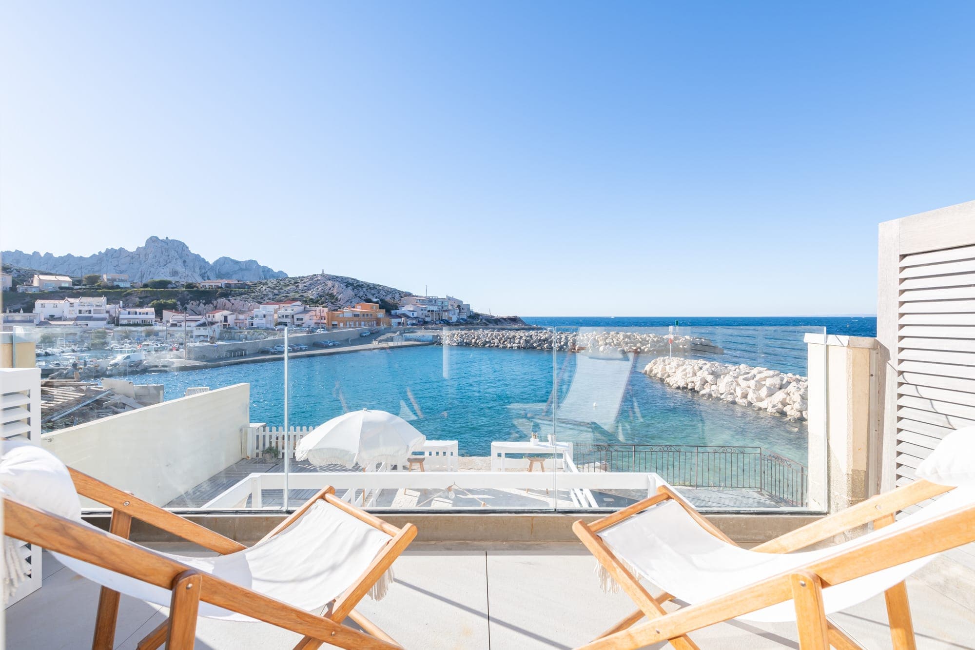 Une terrasse ensoleillée avec deux chaises longues en bois surplombant une baie bleue sereine. La vue comprend une petite marina, un brise-lames rocheux et une colline lointaine avec des maisons. Des parasols blancs et un ciel clair complètent le cadre balnéaire tranquille.
