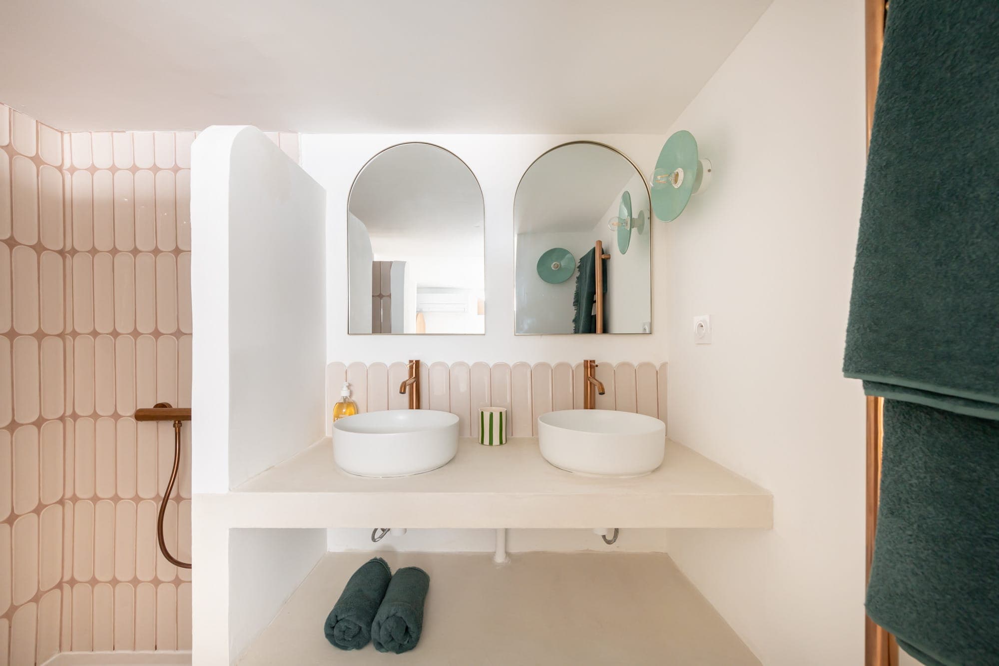 Salle de bain moderne avec deux vasques blanches et miroirs ronds sur un plan de travail blanc. Dosseret en carrelage rose et allongé, appliques vertes, serviettes sous le comptoir et douche à l'italienne à gauche avec des accessoires marron.