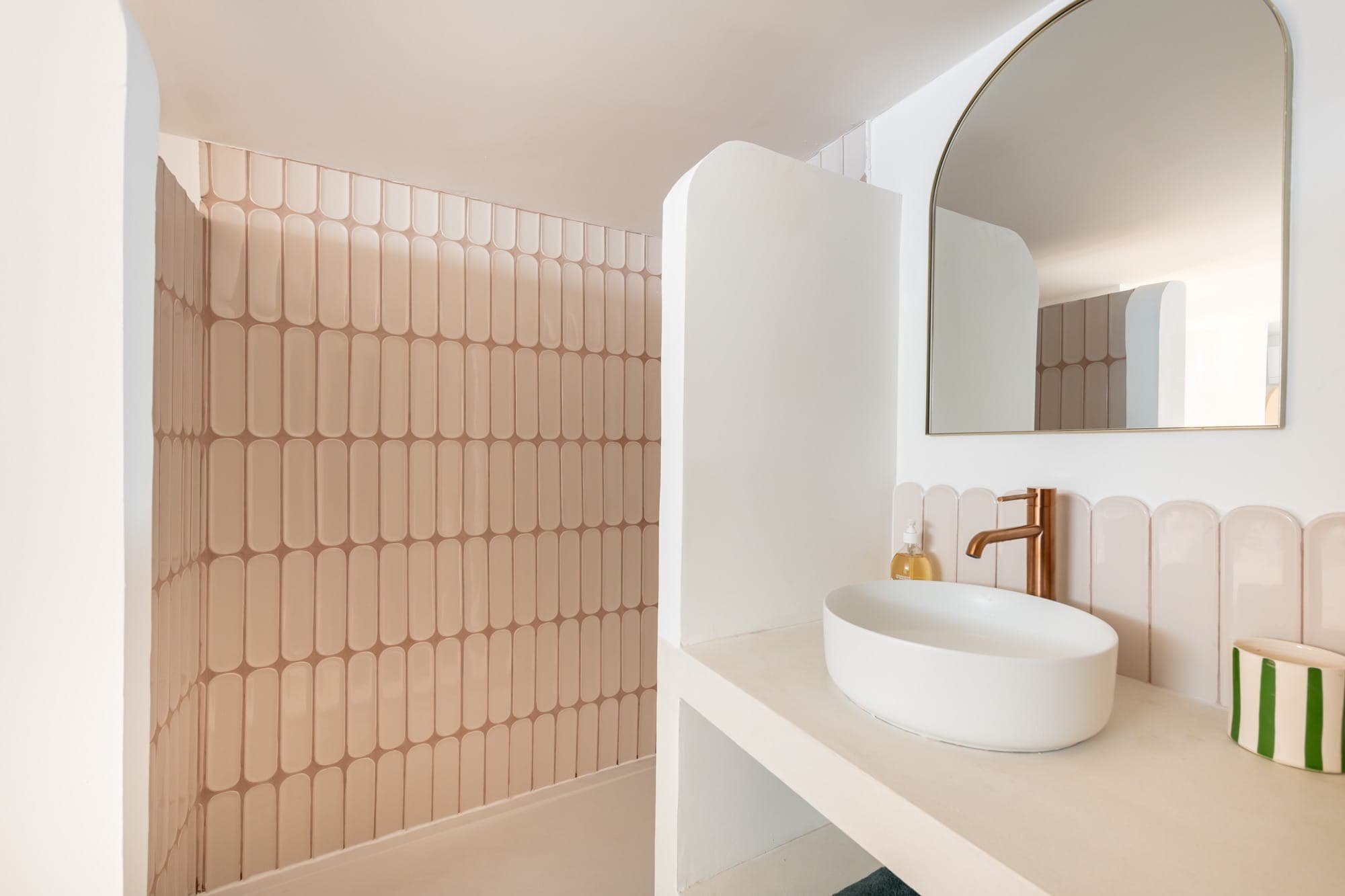 Une salle de bain minimaliste avec une douche à l'italienne avec des carreaux beiges verticaux, un comptoir blanc avec un lavabo rond, un miroir incurvé au-dessus et un robinet moderne en finition cuivre.