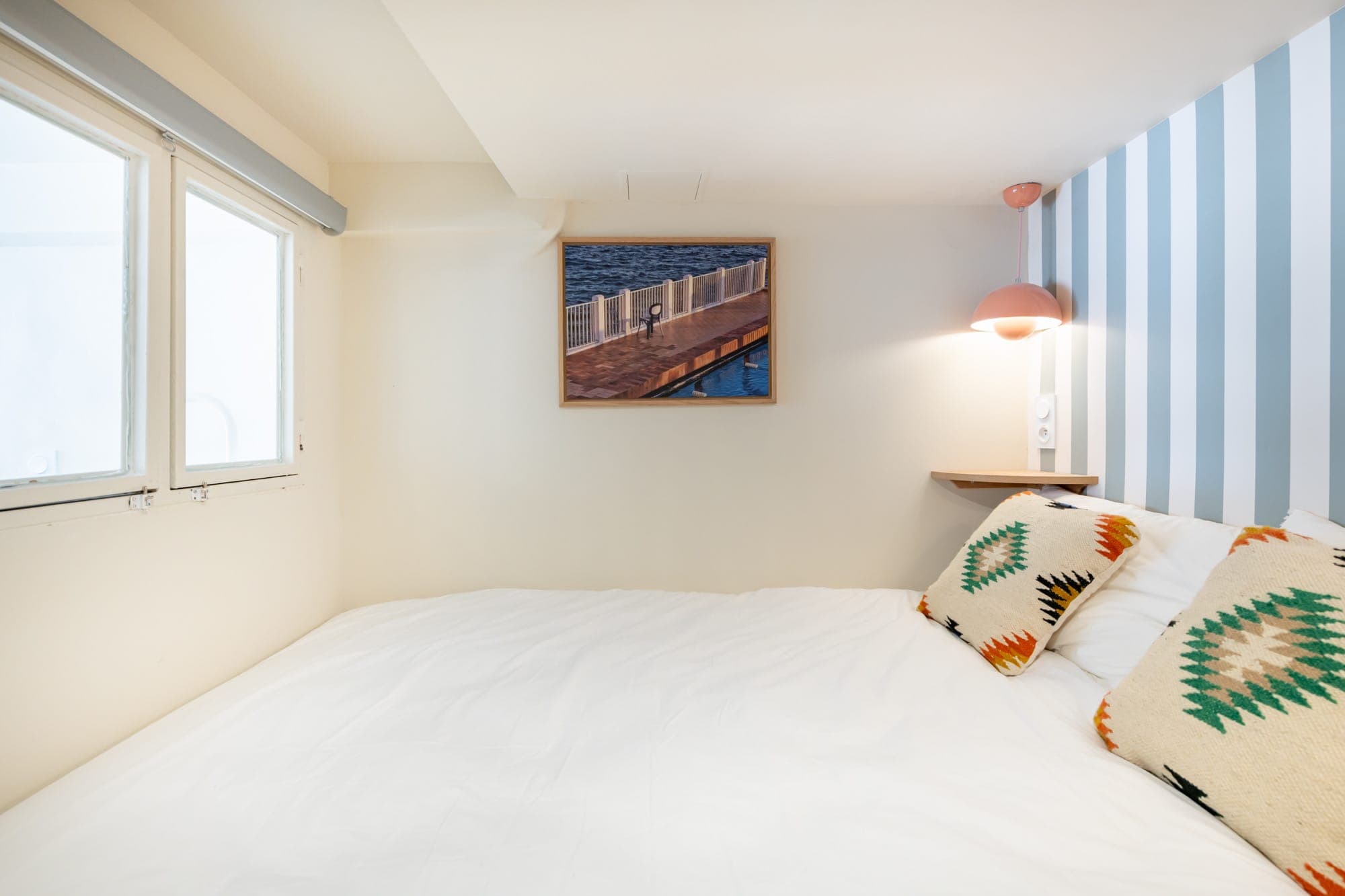 Une chambre confortable comprend un lit blanc avec des coussins géométriques colorés, un mur d'accent rayé avec une lampe montée et un tableau représentant une jetée au-dessus du lit. La lumière naturelle pénètre par les fenêtres voisines, créant une atmosphère lumineuse.