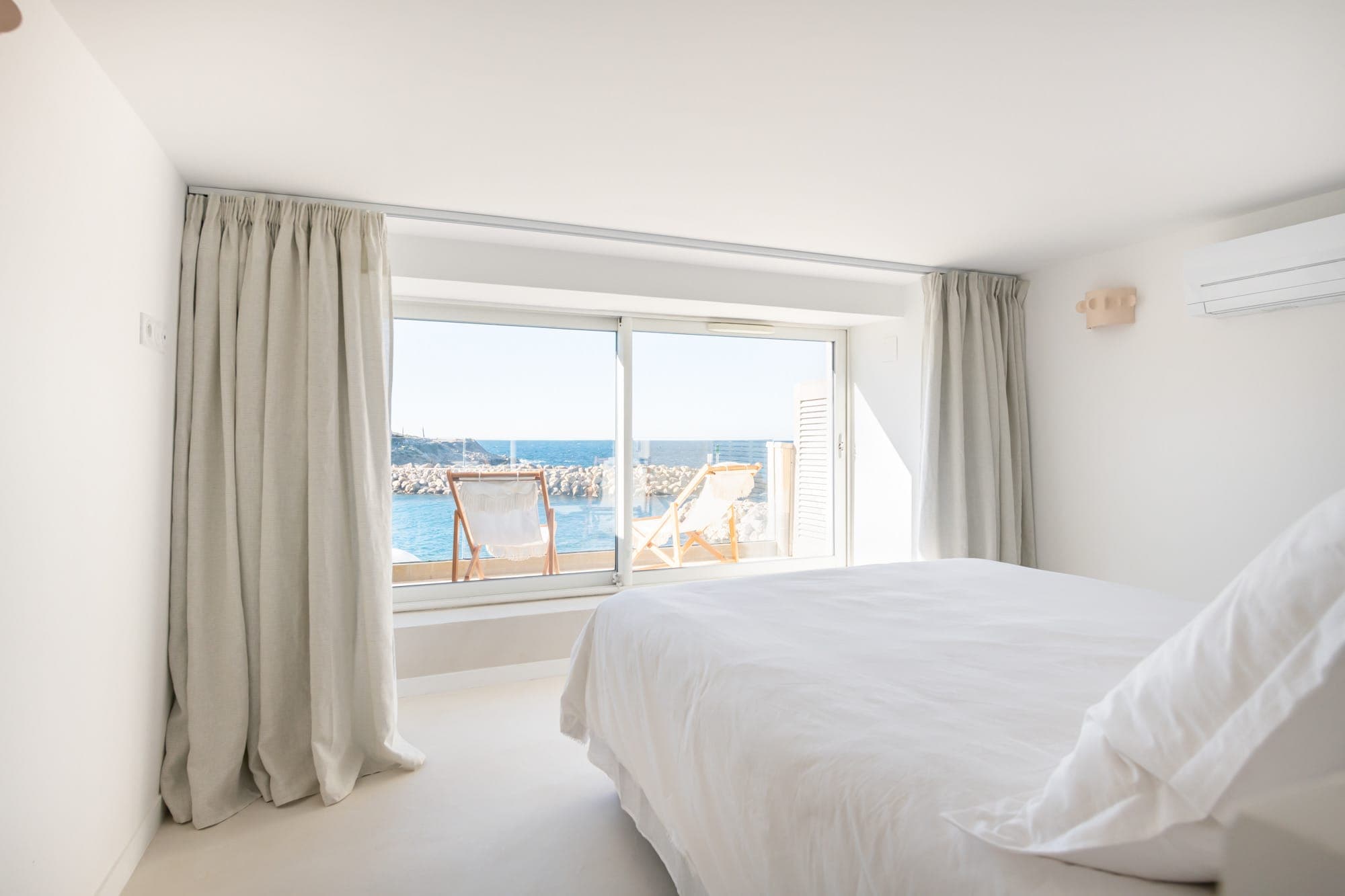 Une chambre lumineuse et minimaliste avec une grande fenêtre donnant sur l'océan. La chambre dispose d'un lit bien fait, de rideaux blancs et de deux chaises de plage sur le petit balcon extérieur, offrant une vue sereine sur la mer et le littoral.