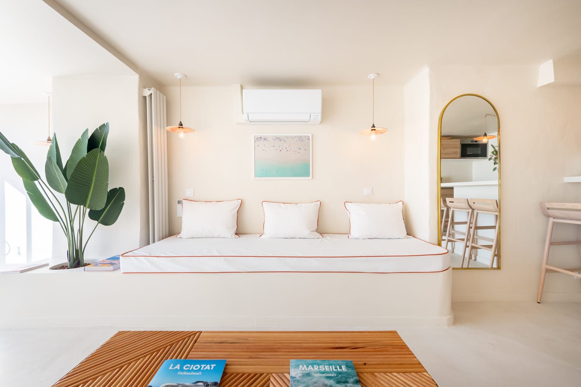 Une pièce lumineuse et minimaliste avec un canapé blanc orné de coussins, une grande plante, un miroir mural et des objets de décoration. Une table basse en bois au premier plan avec des livres. Les tons neutres créent une atmosphère sereine.