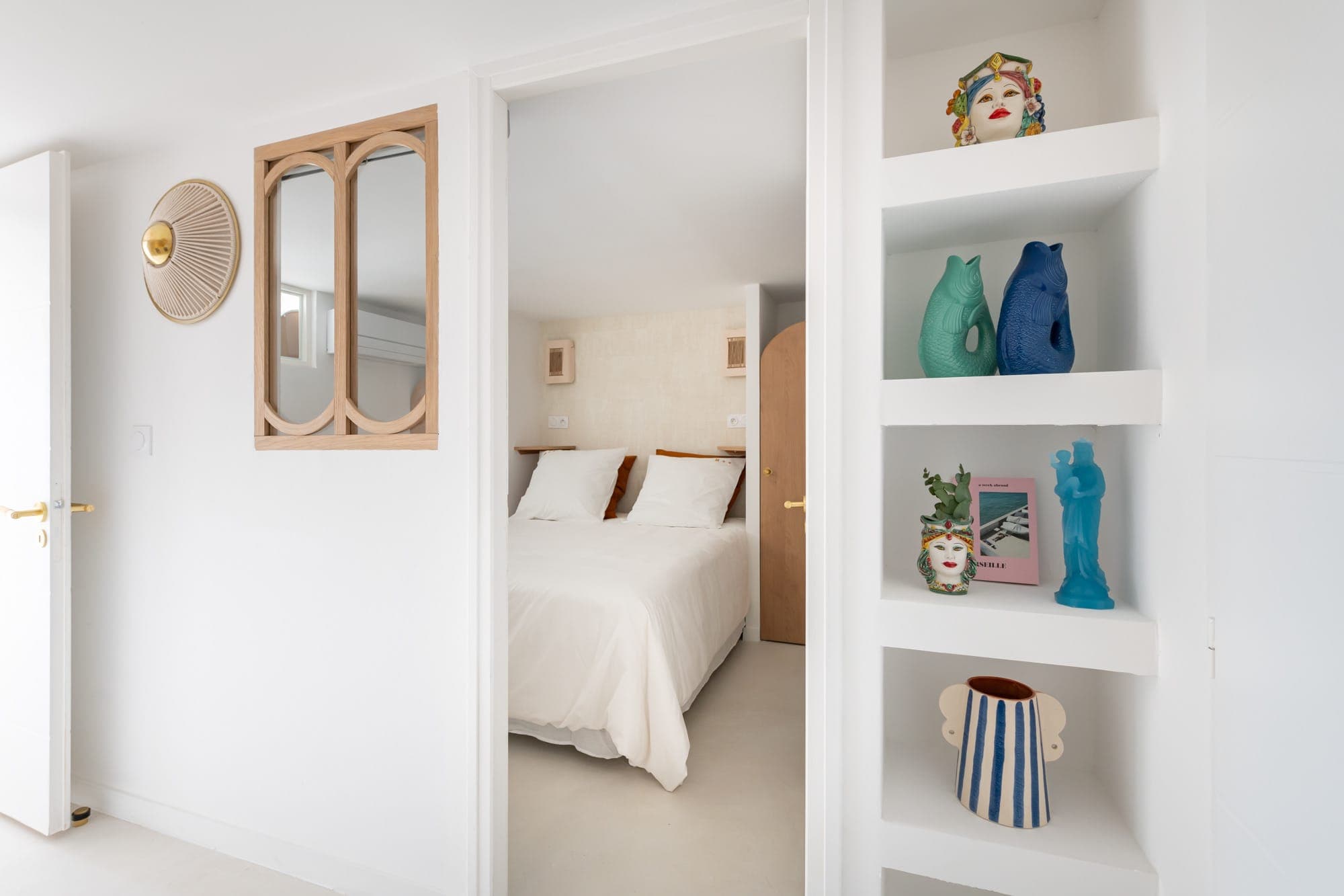 Une chambre lumineuse et minimaliste avec un lit bien fait et des draps blancs. Des étagères ouvertes présentent des poteries décoratives, notamment des vases et une sculpture de visage. Les murs et le sol sont blancs, renforçant la sensation d'espace de la pièce.