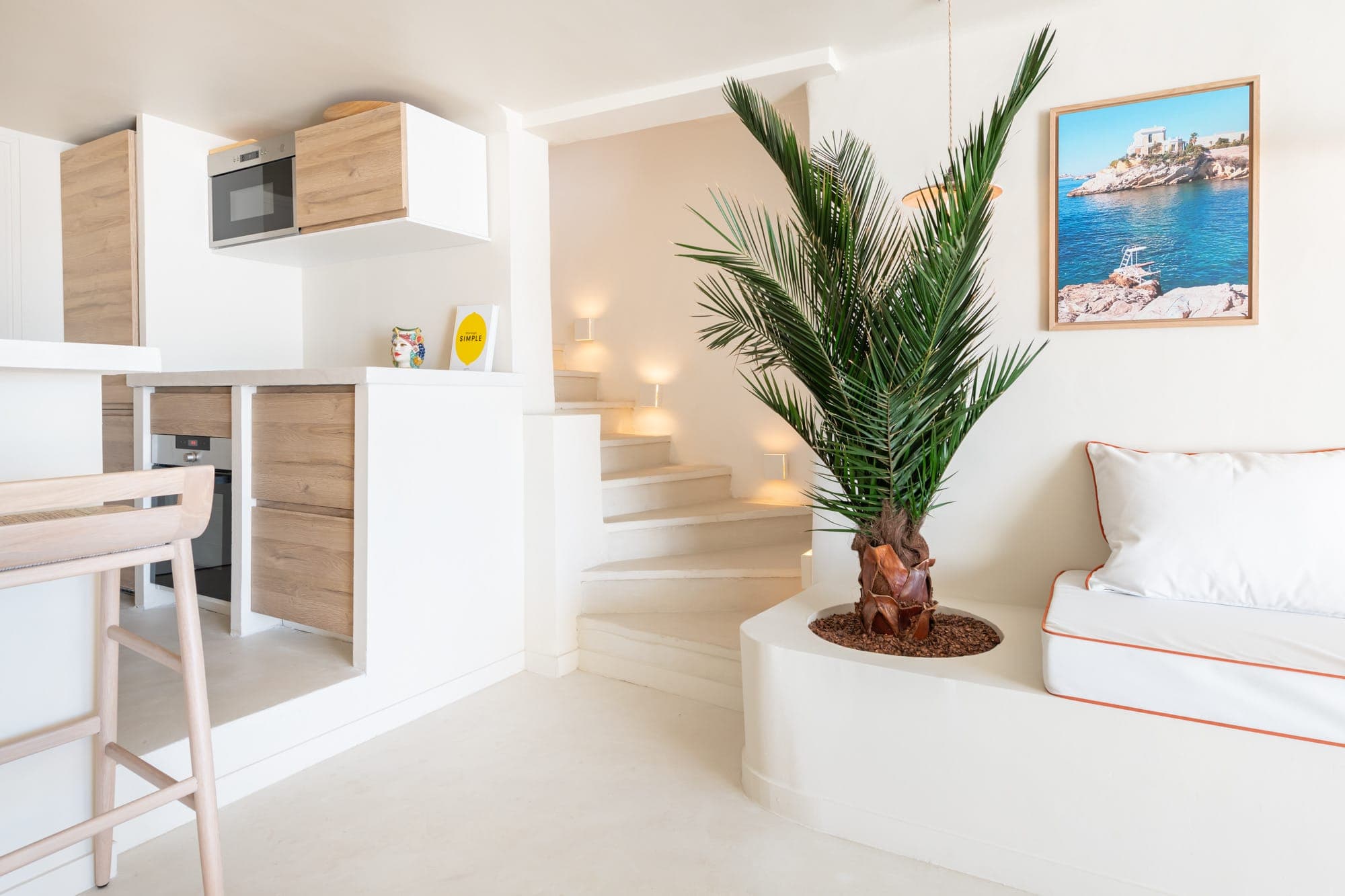 Intérieur blanc moderne avec une petite cuisine aux accents boisés, un palmier en pot et un coin salon blanc avec passepoil orange. Un tableau côtier encadré est accroché au mur et un escalier mène à un niveau supérieur, doucement éclairé.