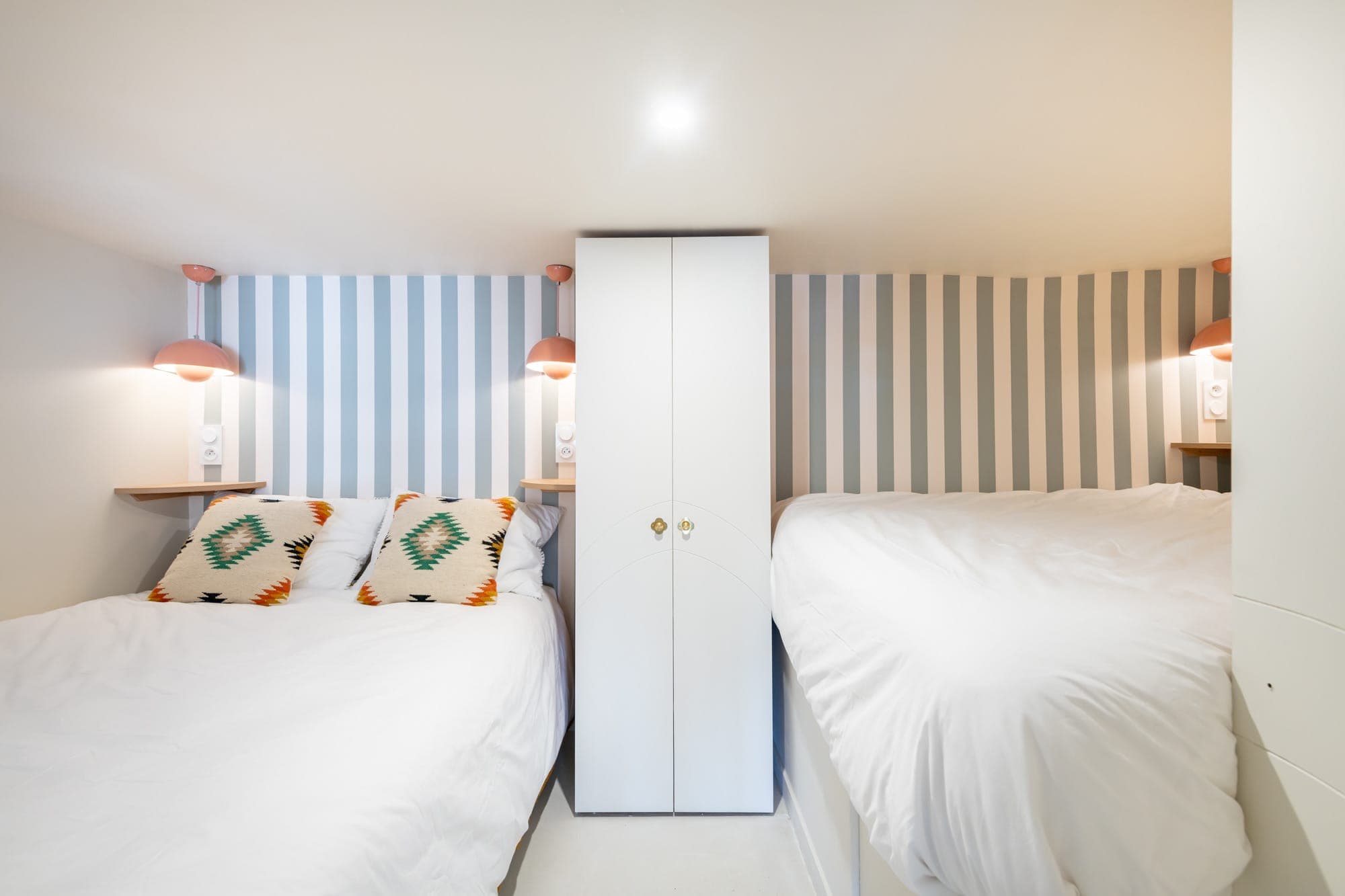 Une chambre confortable avec deux lits simples, chacun avec une literie blanche. Entre les lits se trouve une armoire blanche. Les murs sont décorés de rayures verticales dans des tons bleus et blancs. Des lampes murales et des coussins à motifs ajoutent une touche de couleur.