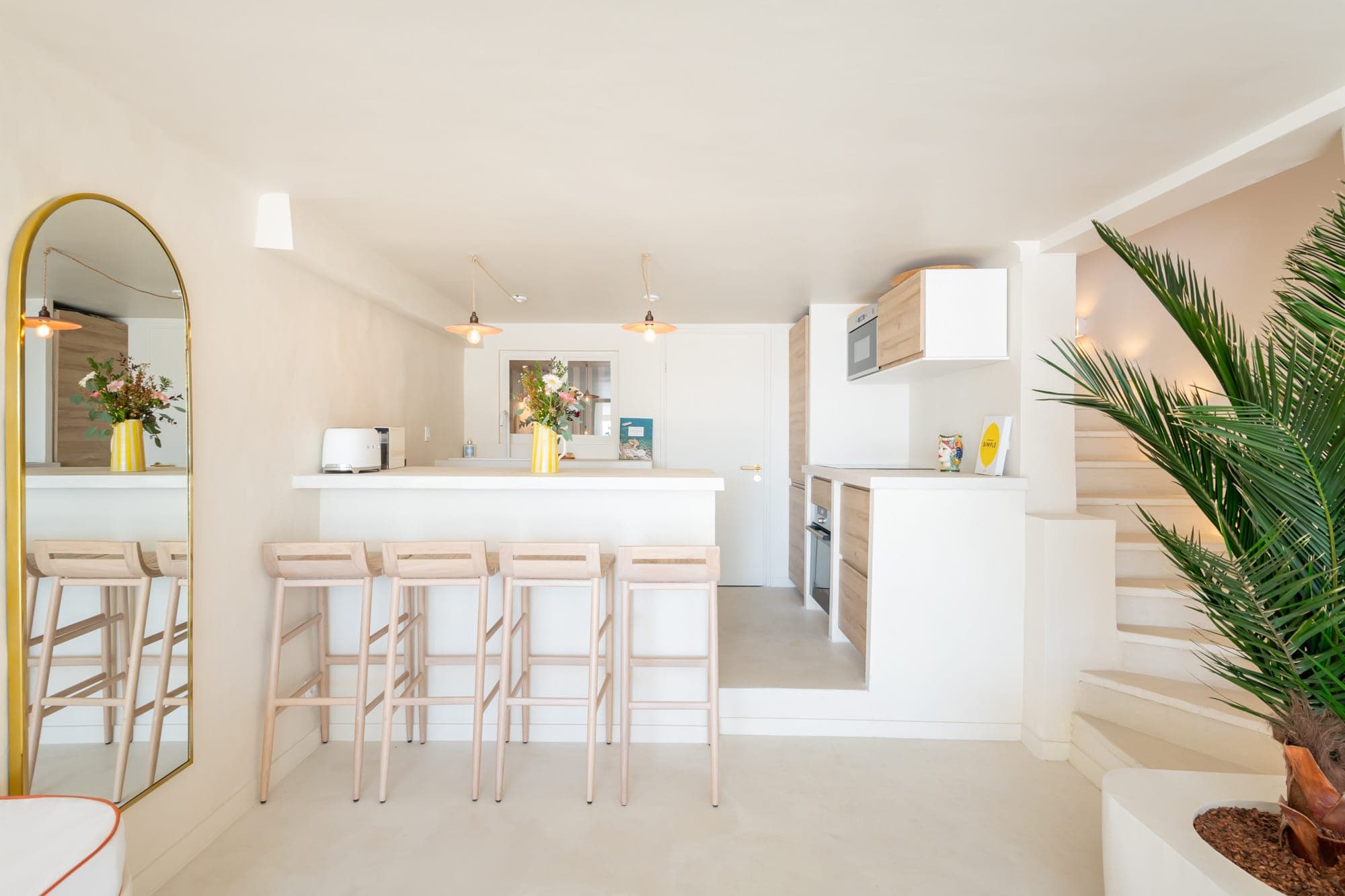 Cuisine lumineuse et moderne avec un long comptoir blanc et quatre tabourets de bar en bois. Un miroir avec un cadre doré est adossé à un mur à gauche. Une plante en pot se trouve à droite et des escaliers mènent vers le haut. La pièce est décorée de fleurs fraîches et de luminaires suspendus.