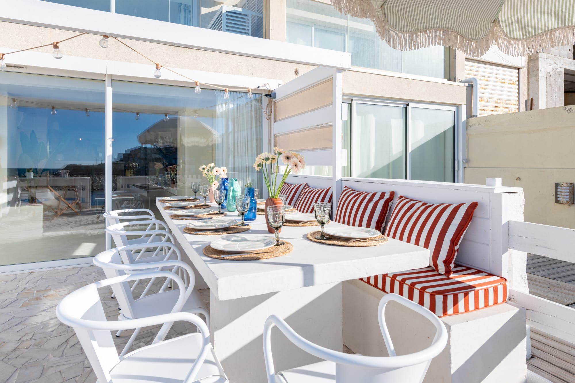 Un espace repas extérieur moderne avec une longue table blanche, des chaises blanches et des bancs rembourrés à rayures rouges. La table est dressée avec des assiettes, des verres et des vases de fleurs. Des portes coulissantes en verre et des guirlandes lumineuses blanches sont visibles en arrière-plan.