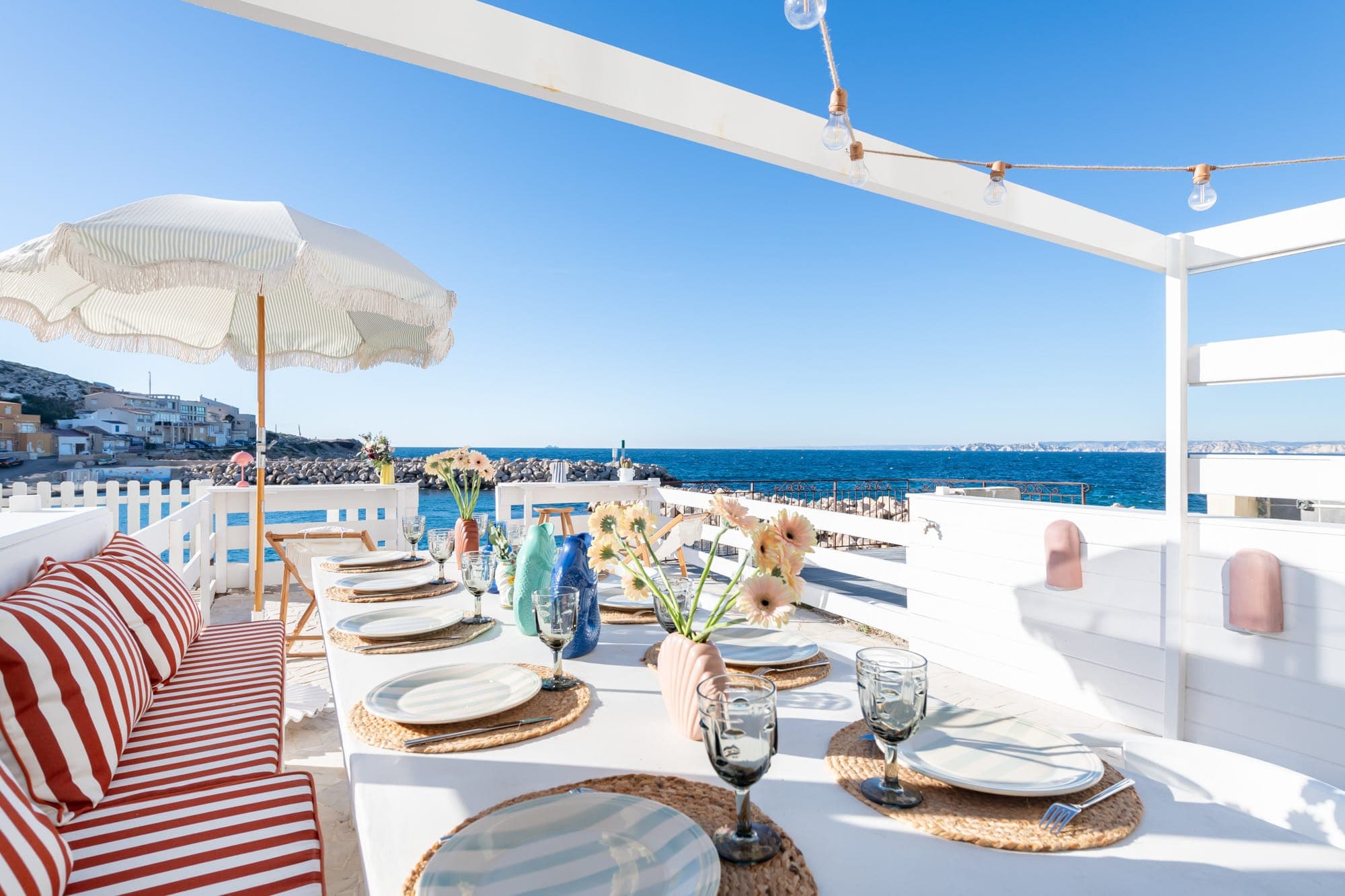 A seaside terrace set for dining with wicker placemats, glassware, and floral arrangements. Striped cushions adorn the bench, and a white umbrella provides shade. The blue sea and clear sky create a serene backdrop.