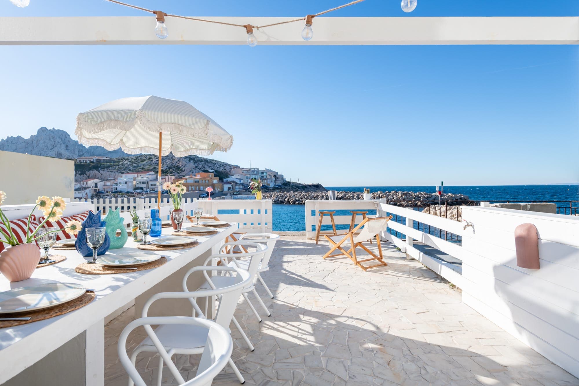 Espace repas extérieur en bord de mer avec mobilier blanc et parasol. Une longue table est dressée avec des assiettes, des verres et des fleurs. En arrière-plan, on aperçoit des falaises rocheuses, un village et l'océan sous un ciel bleu clair.