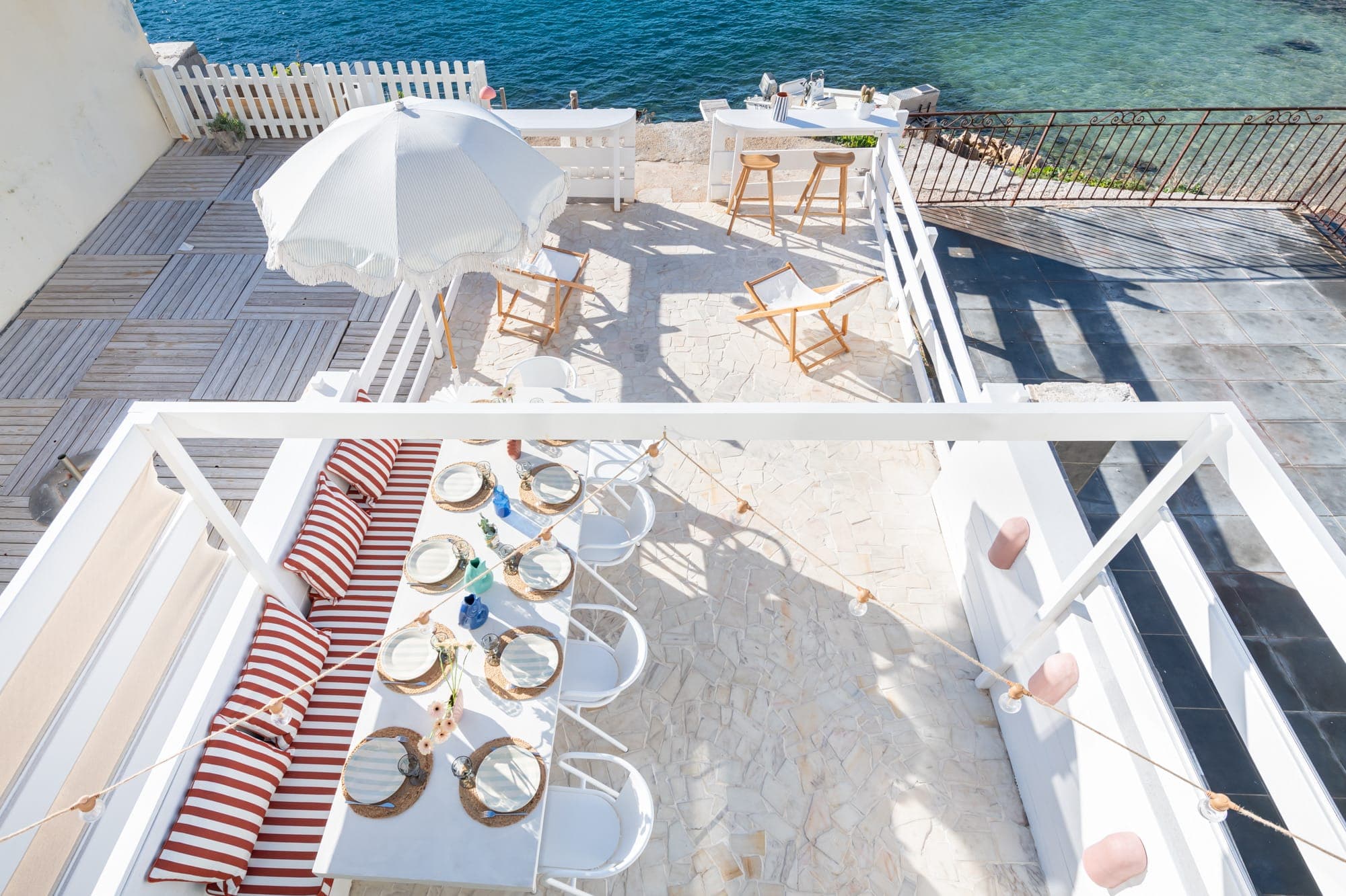 Vue aérienne d'une terrasse ensoleillée en bord de mer avec une table à manger dressée pour un repas. Il y a des coussins à rayures rouges, des chaises blanches, un grand parasol et des chaises longues en bois. L'océan est visible en arrière-plan, avec une eau bleu clair.