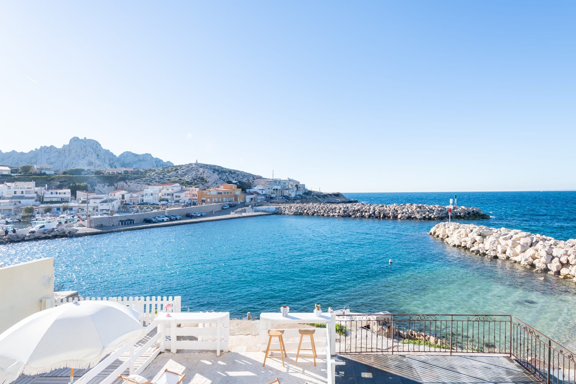 Vue ensoleillée d'une petite ville côtière. Une eau bleue claire remplit une baie tranquille entourée de jetées rocheuses. Des bâtiments blancs bordent le rivage et quelques chaises sont installées sur un balcon au premier plan, surplombant l'océan.