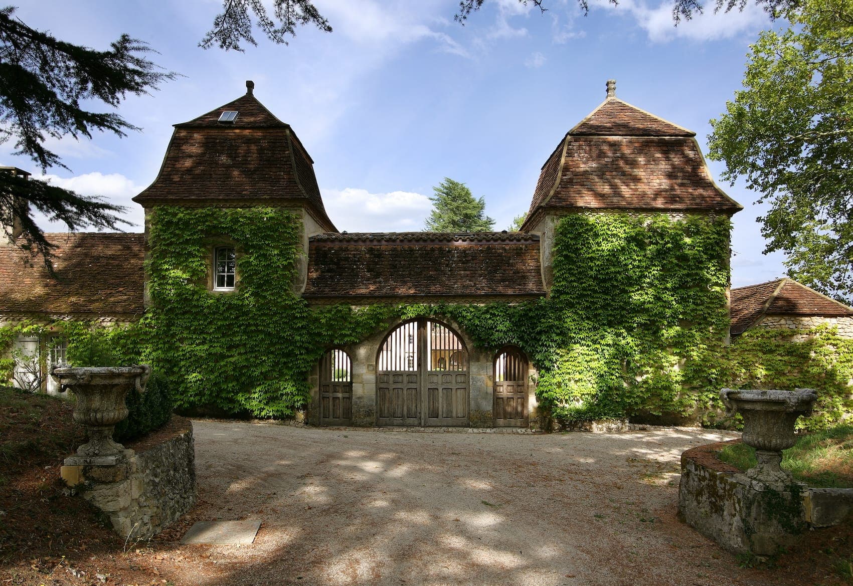 Un bâtiment rustique en pierre recouvert de lierre, avec deux tours et une porte centrale en bois, sur fond de ciel bleu clair. Le bâtiment est entouré d'arbres et est accessible par un chemin de gravier.