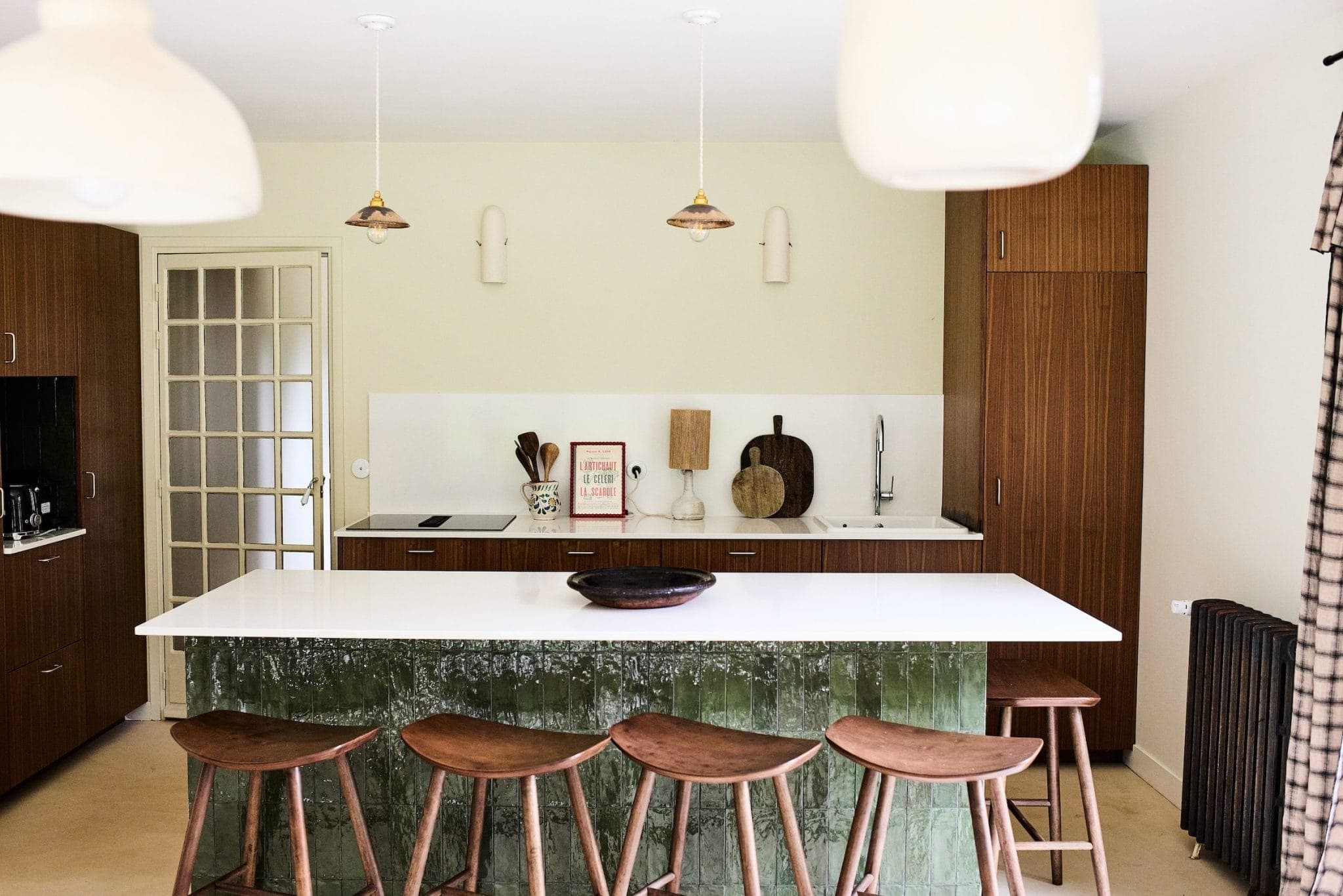 A modern kitchen with wood cabinets and a large white island. Five wooden stools line the island. Pendant lights illuminate the space. A glass door opens to the side, while kitchen utensils and decorative objects adorn the countertop.