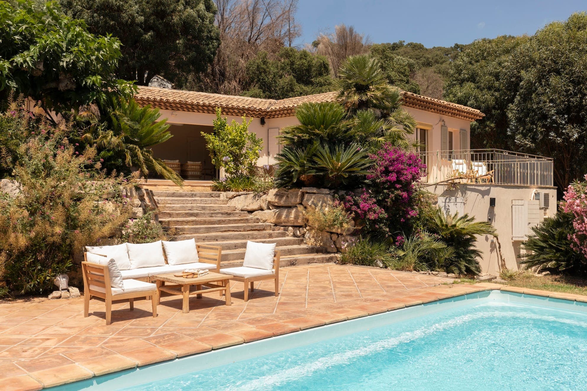 A serene villa with a terracotta roof surrounded by lush greenery and vibrant flowers. It features an outdoor seating area and a swimming pool with clear blue water. Stone steps lead to the villa, set against a backdrop of trees and a clear sky.