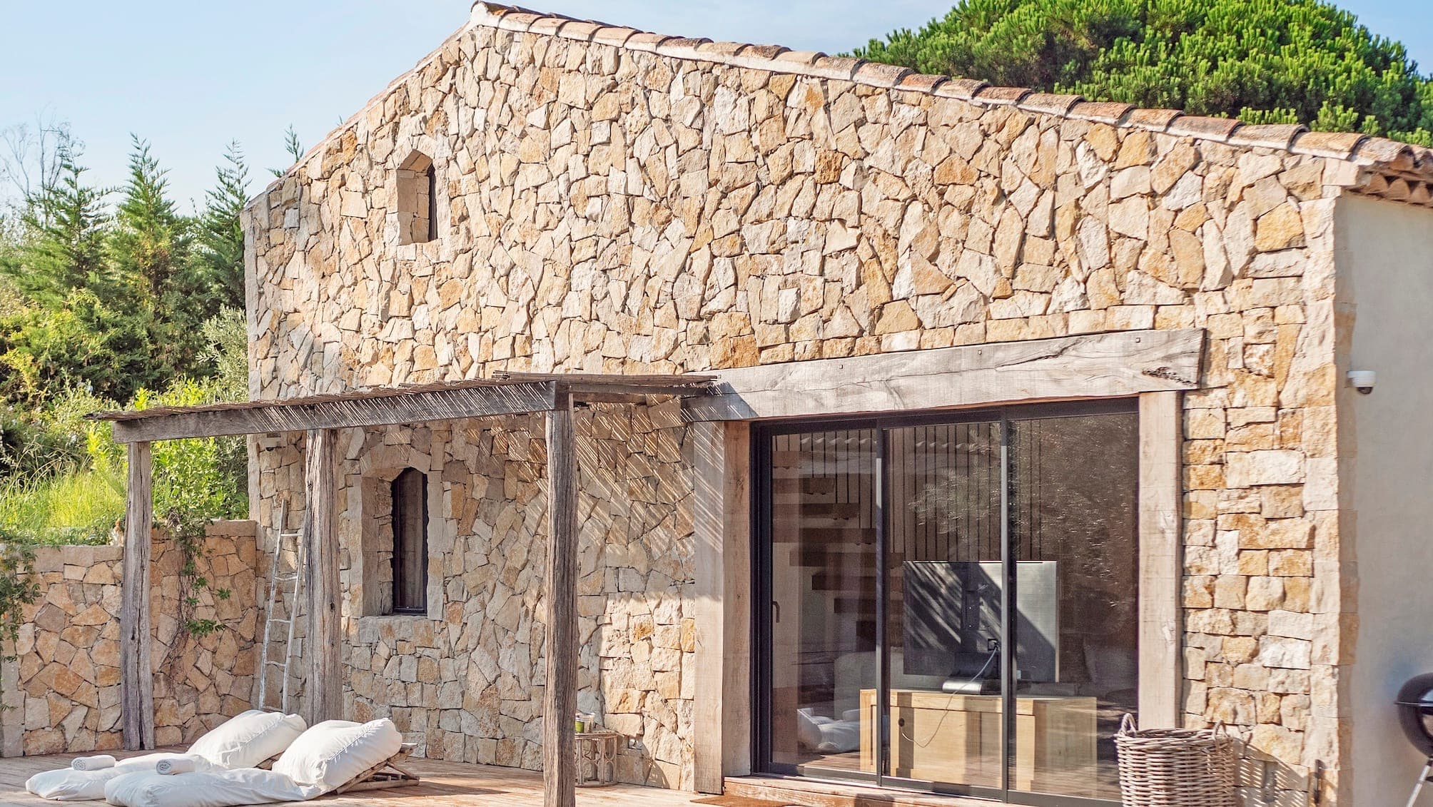 Une maison rustique en pierre avec une pergola en bois sur son porche. De grandes portes coulissantes en verre révèlent un intérieur moderne. Deux coussins blancs sont placés sur la terrasse en bois. Une végétation luxuriante entoure la maison, renforçant son charme naturel.
