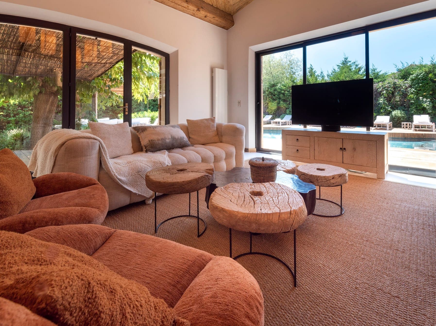 Salon confortable avec un grand canapé d'angle beige avec des coussins moelleux, des tables basses rondes en bois et une télévision à écran plat. Des portes vitrées s'ouvrent sur une cour arrière avec une piscine entourée de verdure. Une décoration aux tons terreux et une ambiance chaleureuse créent un espace accueillant.