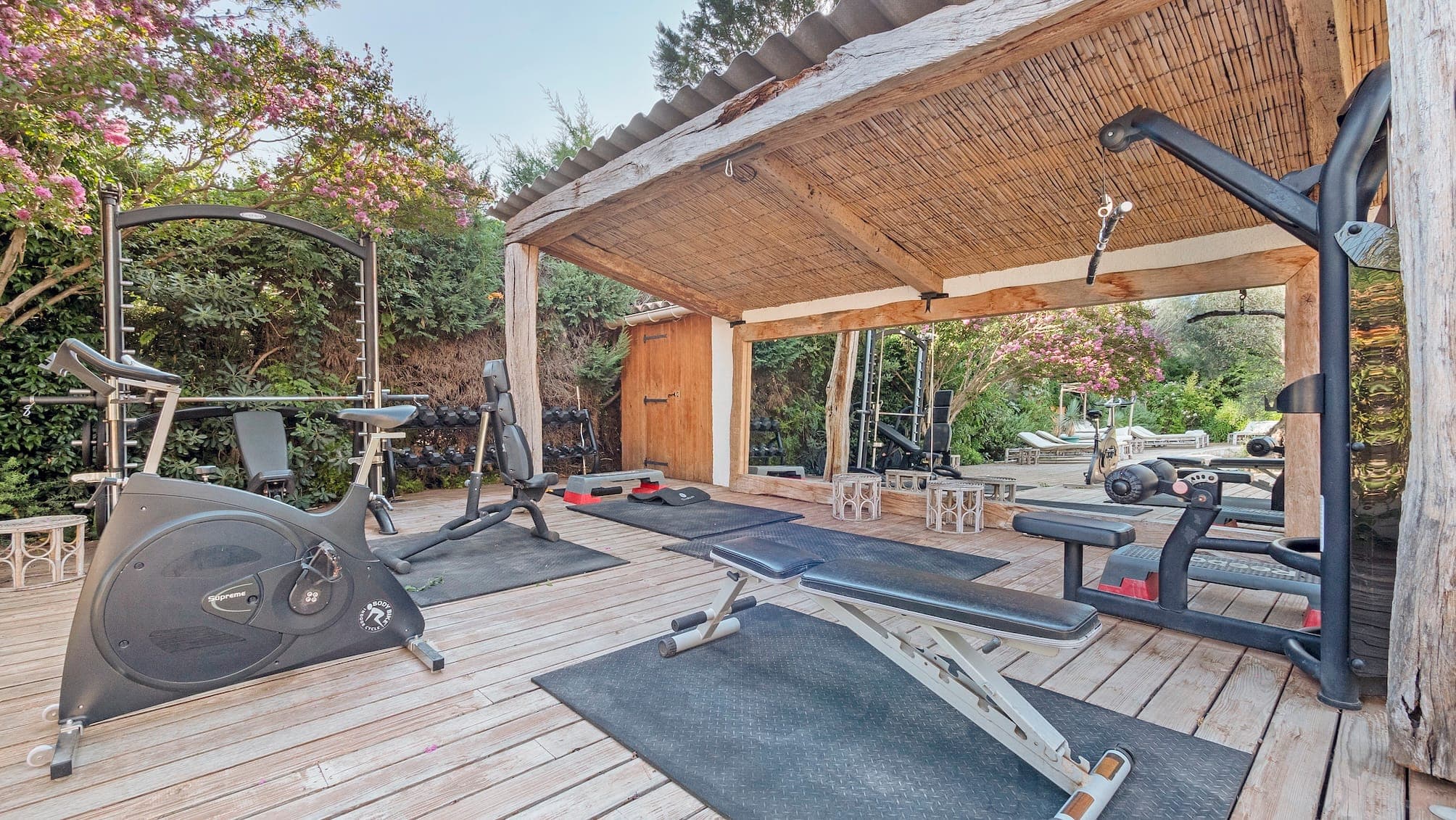 Salle de sport extérieure avec divers équipements de fitness, dont un vélo d'appartement, un banc de musculation et un appareil à câbles. L'espace est aménagé sur une terrasse en bois avec tapis, entourée de verdure et d'arbres à fleurs roses, sous un toit de chaume.