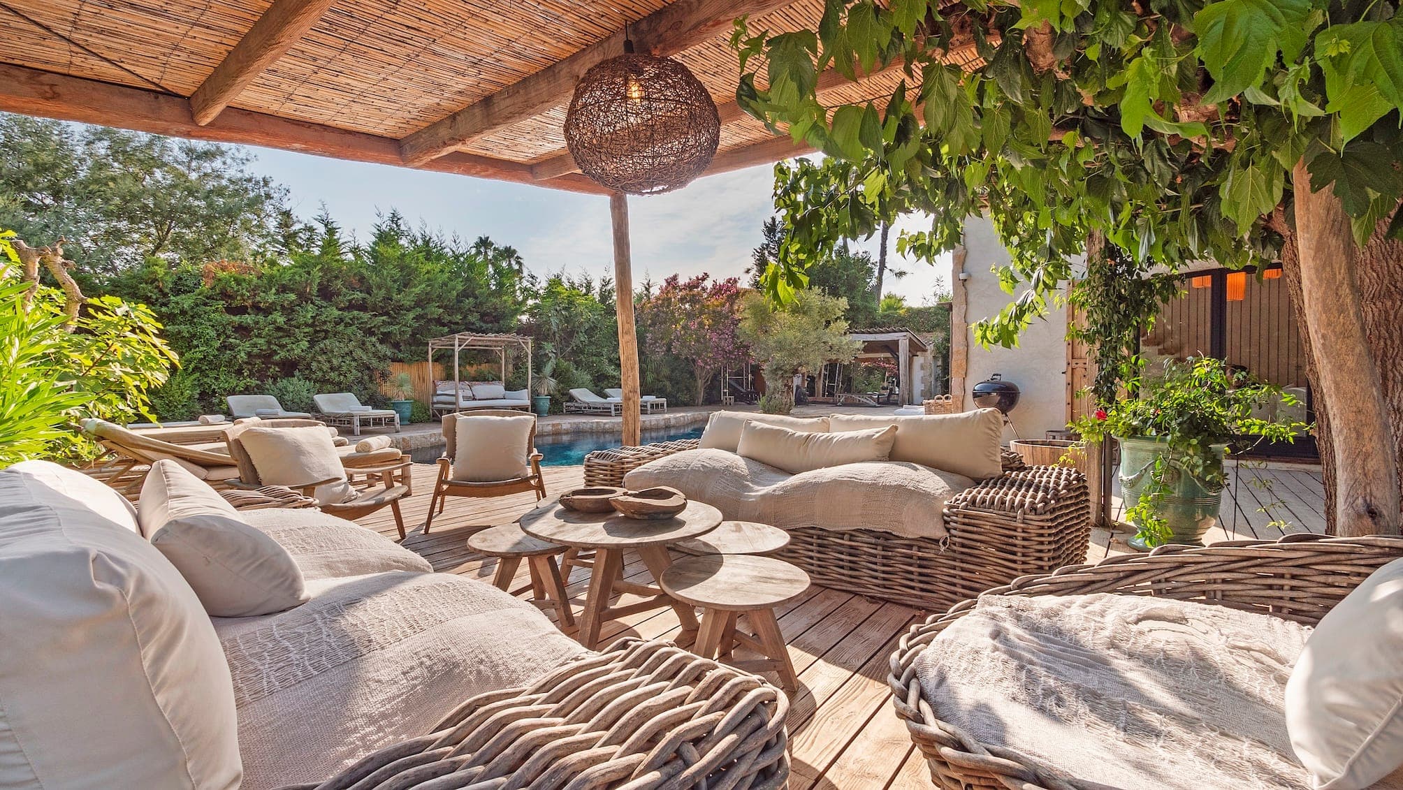 Un espace salon extérieur confortable avec des meubles en osier sous une pergola en bois. Des chaises et des canapés rembourrés entourent une table basse en bois. Une végétation luxuriante et des arbres offrent ombre et intimité, avec un aperçu d'une piscine en arrière-plan.