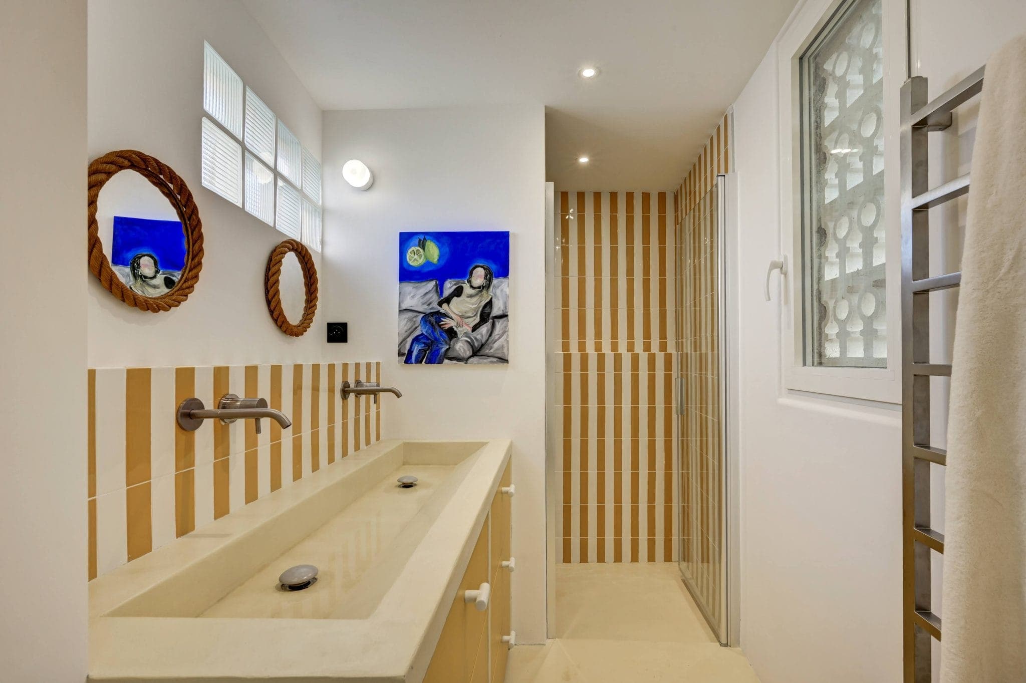 Salle de bains moderne avec un lavabo double et des carreaux à rayures beiges sur le mur. Une grande fenêtre et un tableau représentant une personne portant une veste bleue ajoutent du caractère. Des miroirs circulaires avec des cadres en corde sont montés au-dessus du lavabo et un élégant porte-serviettes se trouve sur la droite.