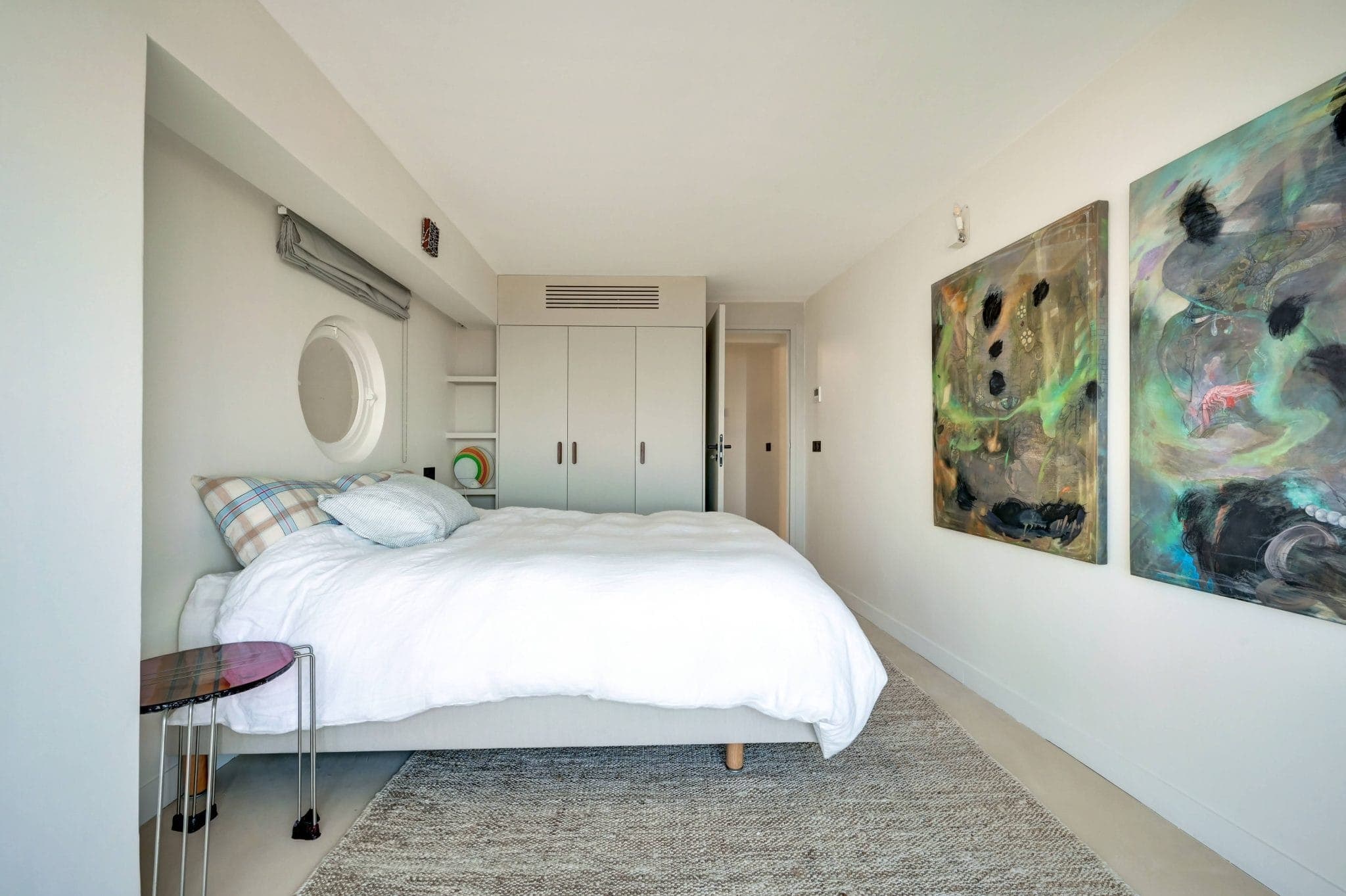 Une chambre minimaliste avec un lit bien fait avec une literie blanche et des oreillers à carreaux. La pièce a des murs clairs, des œuvres d'art abstraites colorées, un miroir rond, une petite table d'appoint et des armoires de rangement intégrées. La lumière naturelle remplit l'espace.