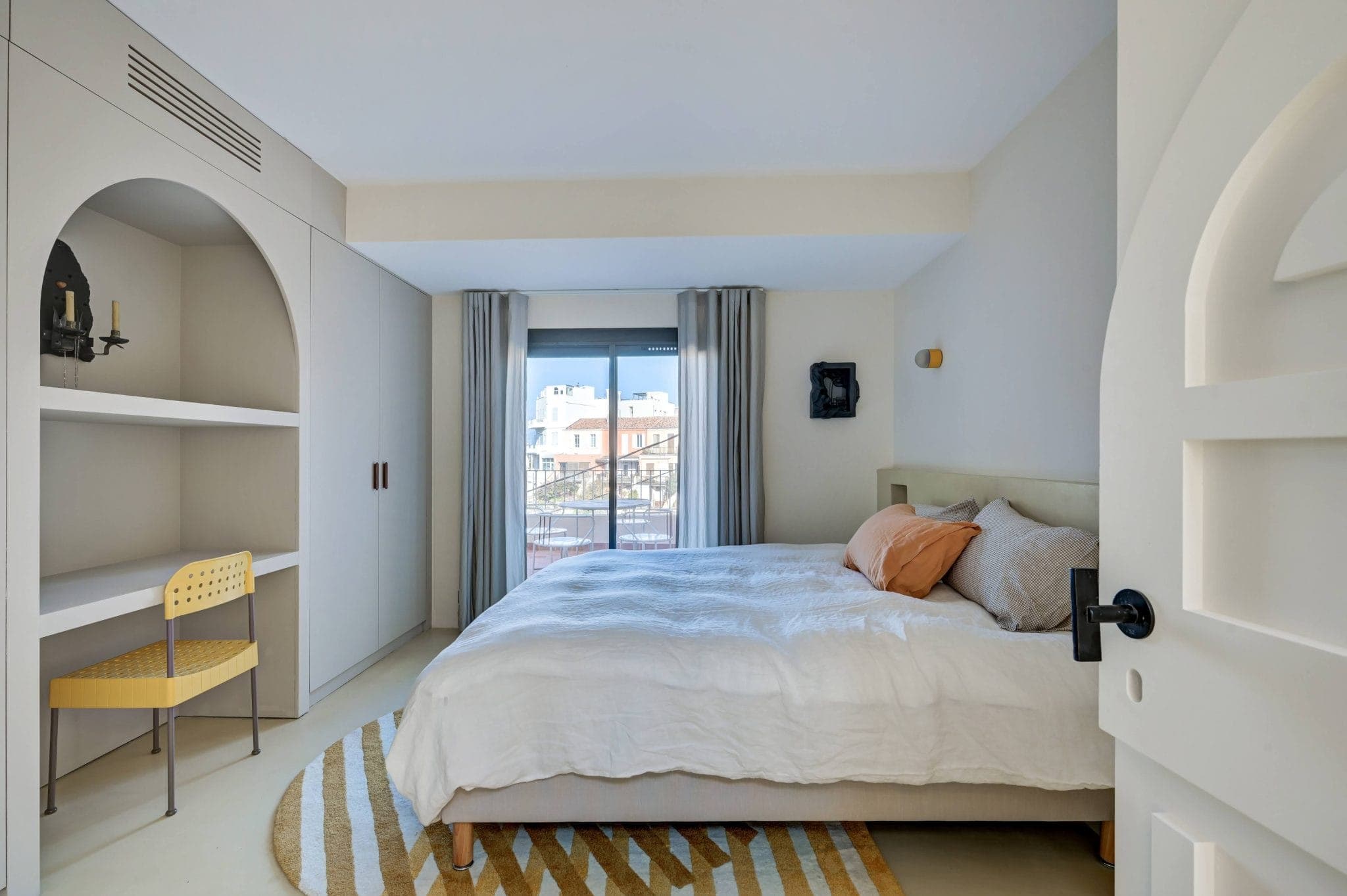 Une chambre bien éclairée avec un lit double, une literie blanche et des oreillers couleur pêche. Il y a une grande fenêtre avec des rideaux gris, une chaise jaune, des étagères intégrées et un tapis décoratif. La pièce est peinte dans des tons beiges doux.