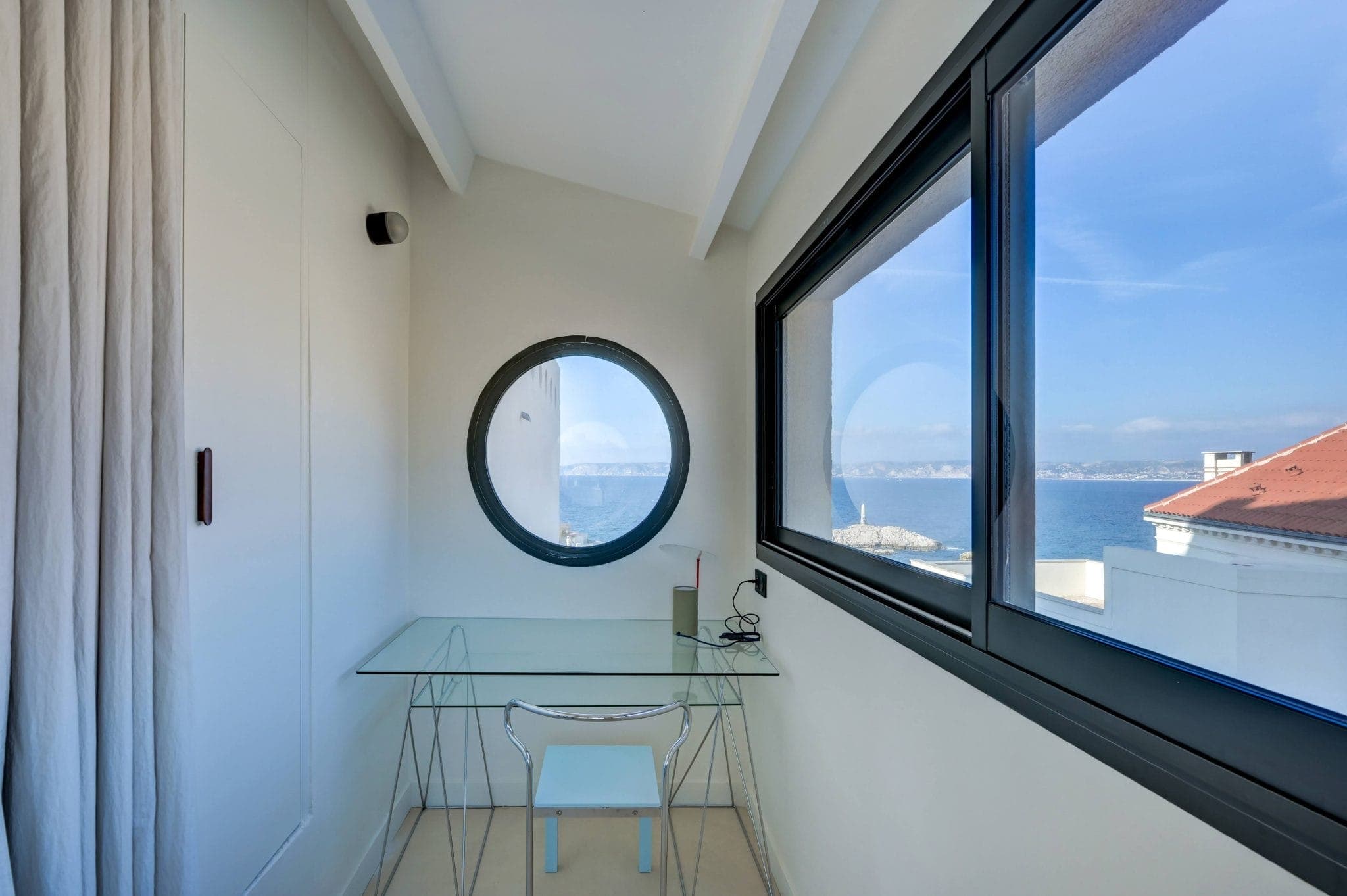 Espace de travail minimaliste avec un bureau en verre et une chaise dans une petite pièce lumineuse. Il y a une fenêtre ronde et une fenêtre rectangulaire avec vue sur l'océan et les toits. Un livre et une lampe sont posés sur le bureau.