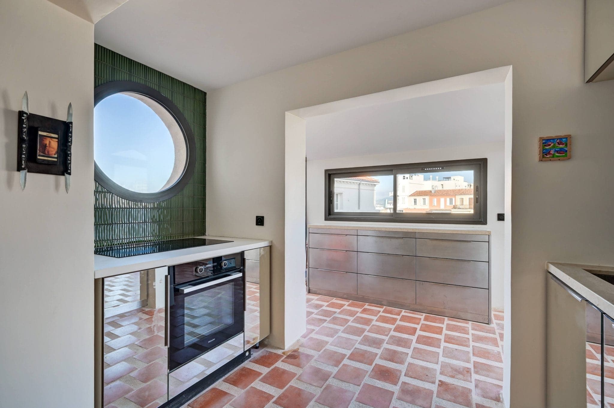 Cuisine moderne avec carrelage en terre cuite, murs blancs et fenêtre circulaire entourée de carreaux verts. Un four encastré est situé sous la fenêtre. Il y a un long comptoir et une petite fenêtre avec vue sur les bâtiments voisins.