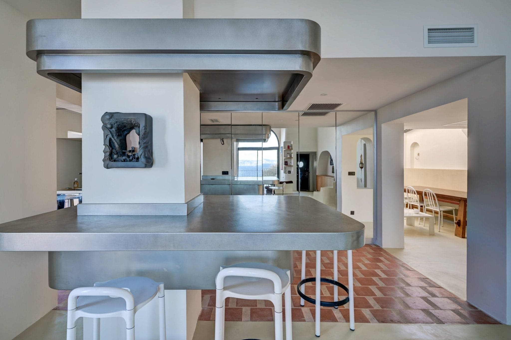 Cuisine moderne avec îlot et cuisinière métalliques, avec une petite sculpture en pierre sur le mur. L'espace est doté de tabourets blancs et d'un sol carrelé à carreaux en terre cuite et blanc. Au fond, il y a un coin repas avec une table en bois et des chaises blanches.