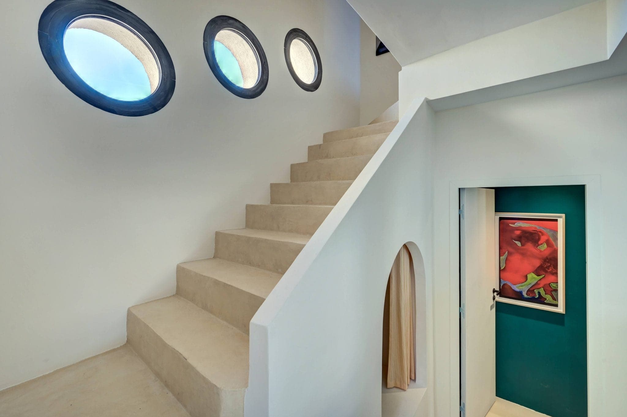 Un intérieur moderne se compose d'un escalier beige lisse avec trois fenêtres circulaires sur le mur. En dessous se trouve un couloir avec une œuvre d'art abstraite colorée sur un mur bleu sarcelle et une petite arche menant à un autre espace.