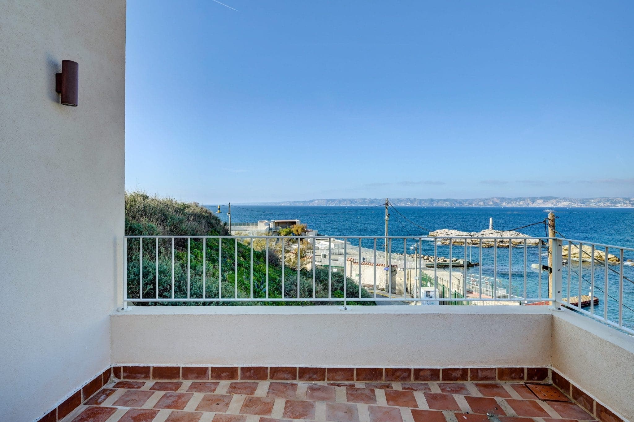 Un balcon en bord de mer avec un sol carrelé et une balustrade blanche offre une vue panoramique sur l'eau bleue de l'océan et les montagnes lointaines sous un ciel clair. Une petite jetée et un phare sont visibles au loin.